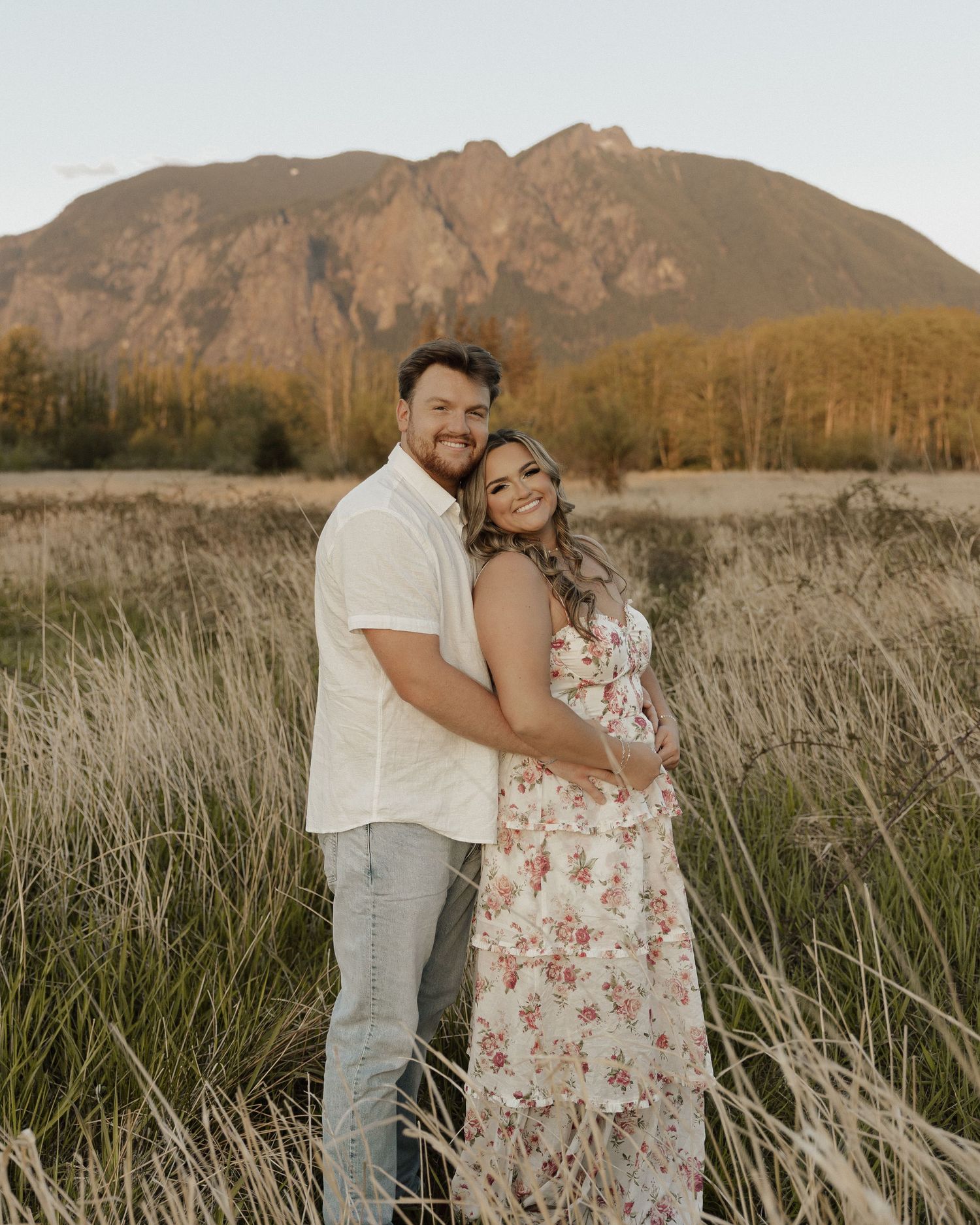 Couple poses together in tall grass with mountain vista during sunset.