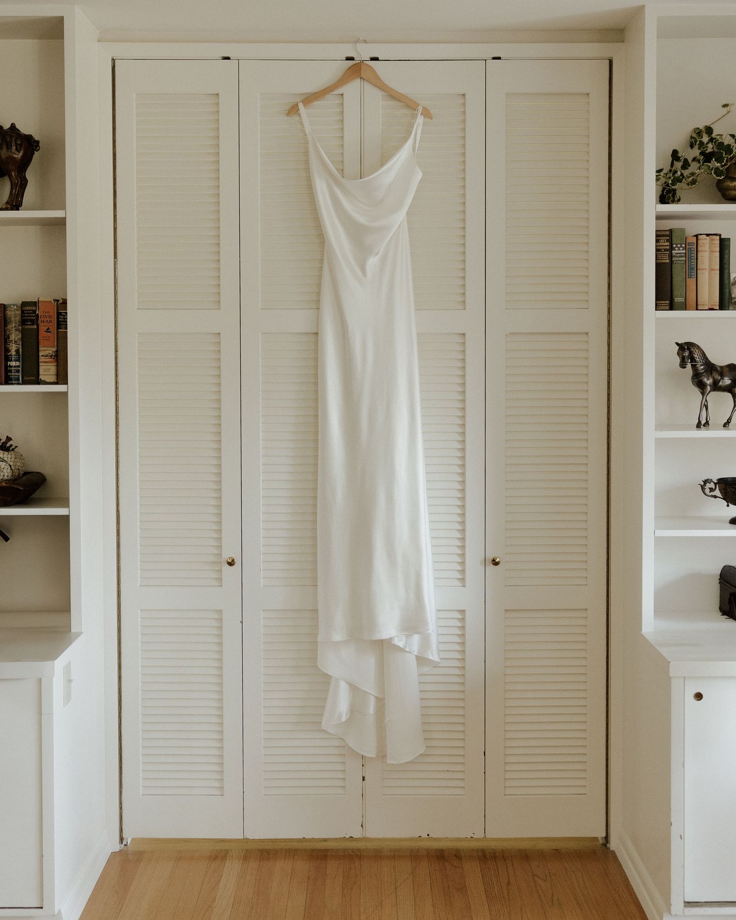 White satin wedding dress hanging on wooden hanger against white louvered closet doors in bright room.