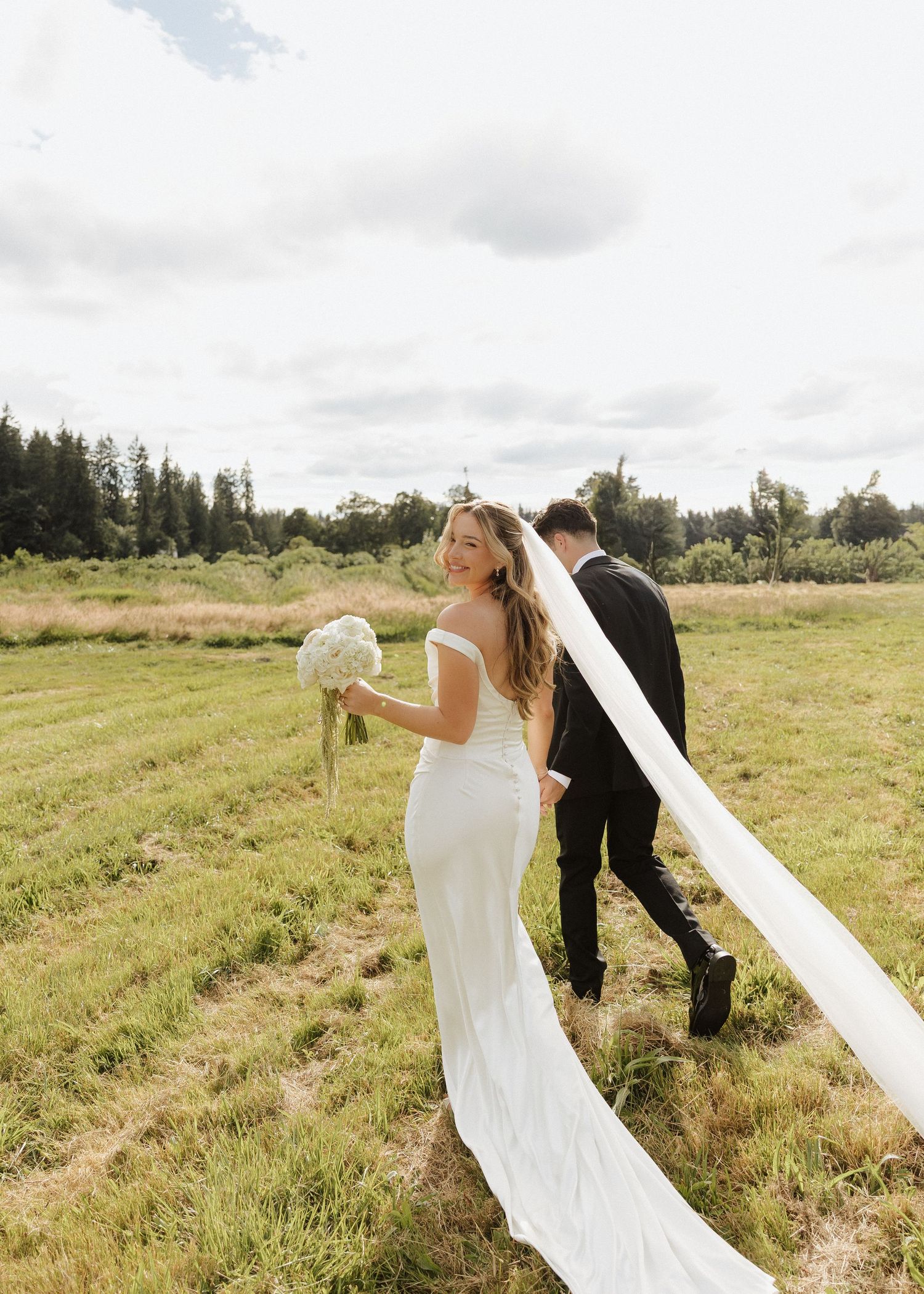 Bride in white gown with long veil walks with groom across lush green field on their wedding day.