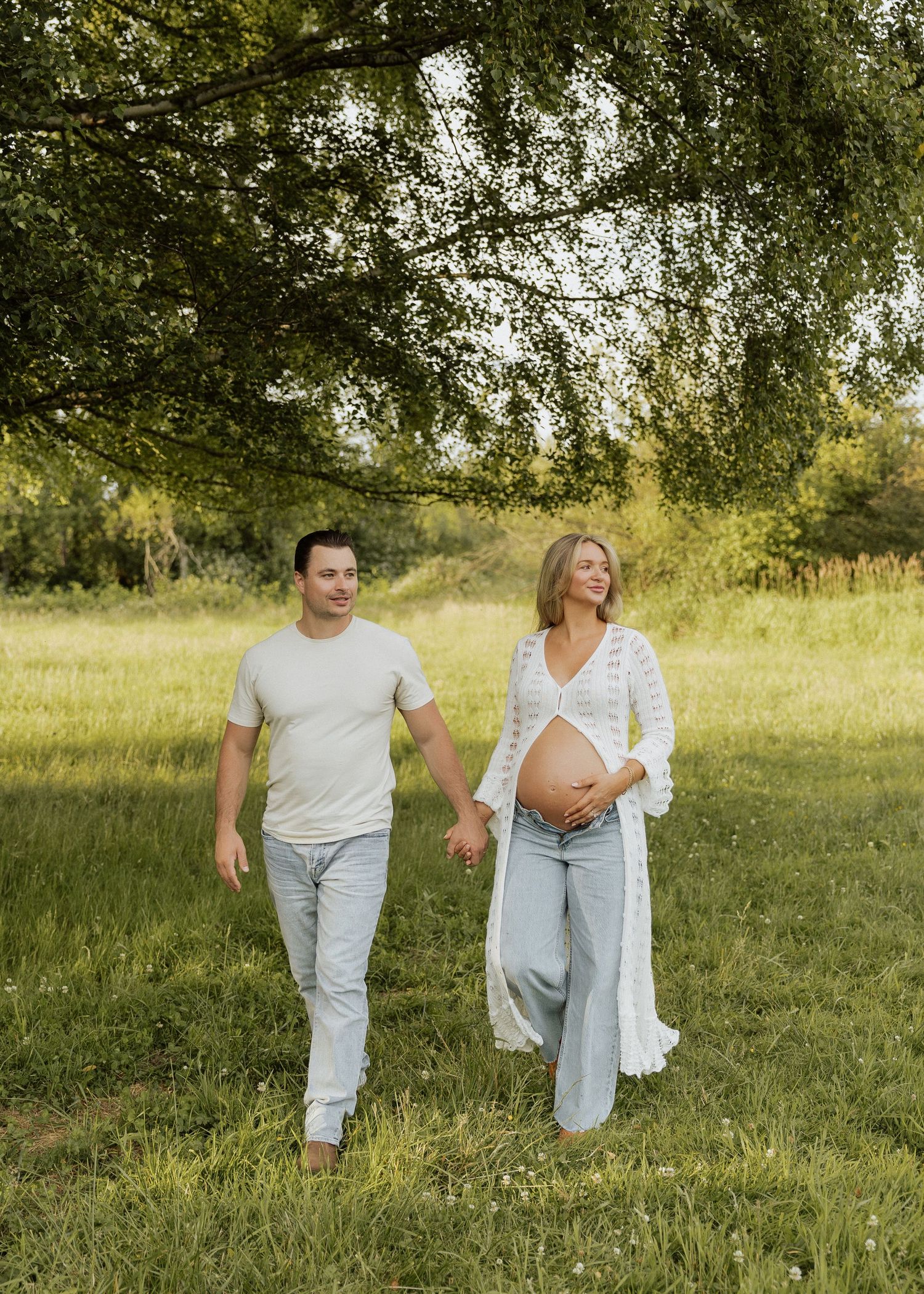 Pregnant couple walking hand-in-hand through a lush green meadow, woman in white lace duster cradling baby bump.