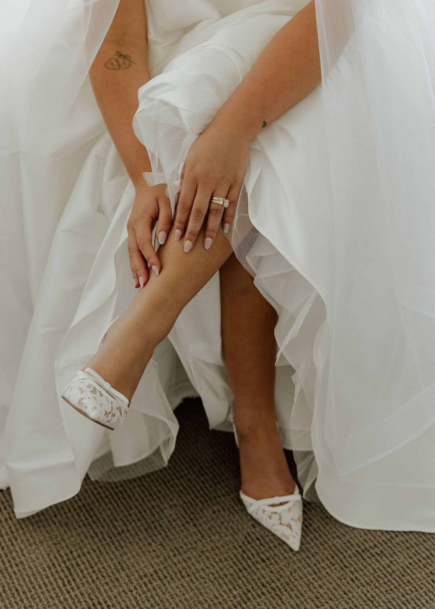 Bride in white wedding gown wearing lace heels and diamond rings, gently holding her ankle in an elegant pose.