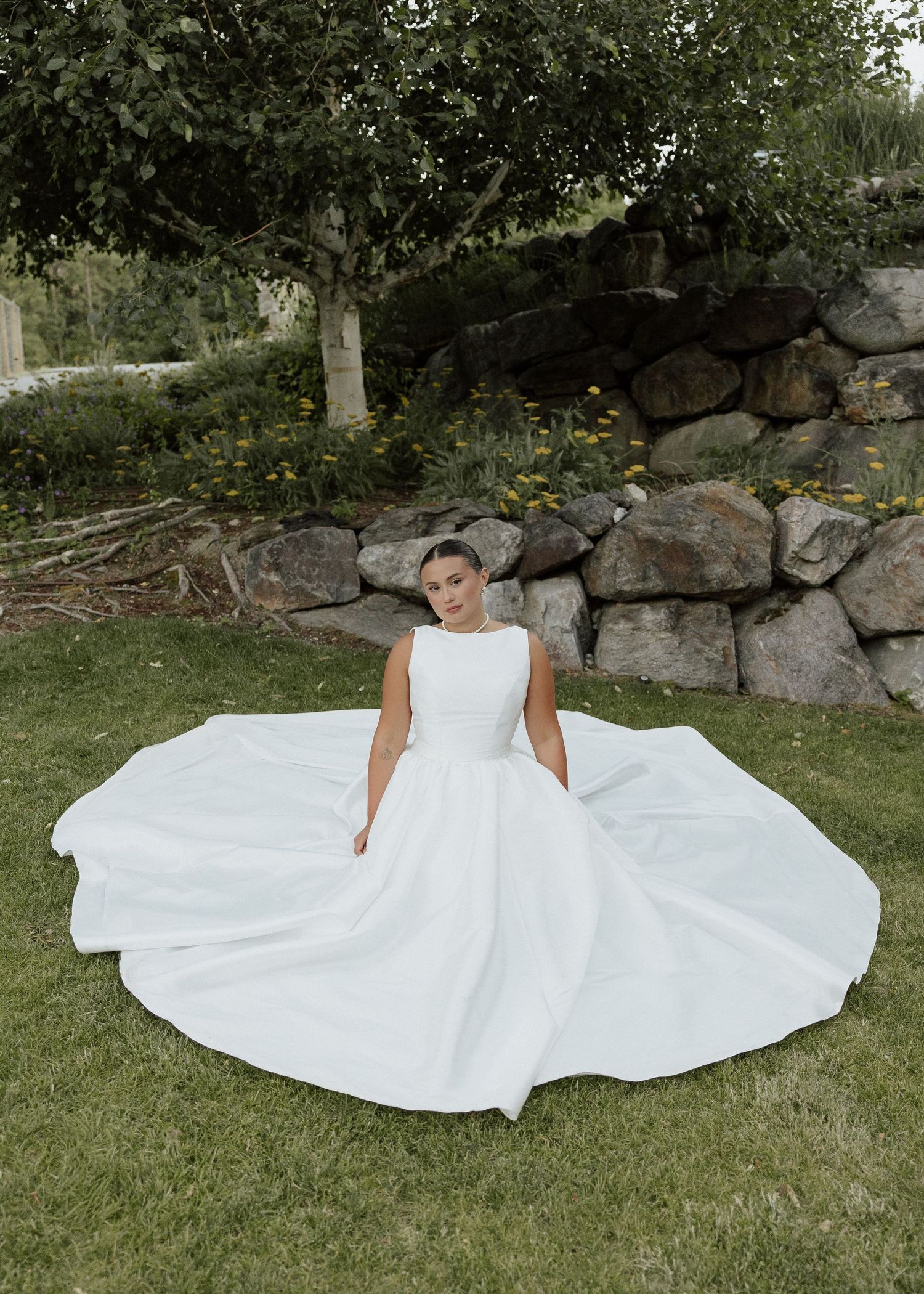 Bride in elegant white ballgown spreading full skirt while sitting on lawn near stone wall.