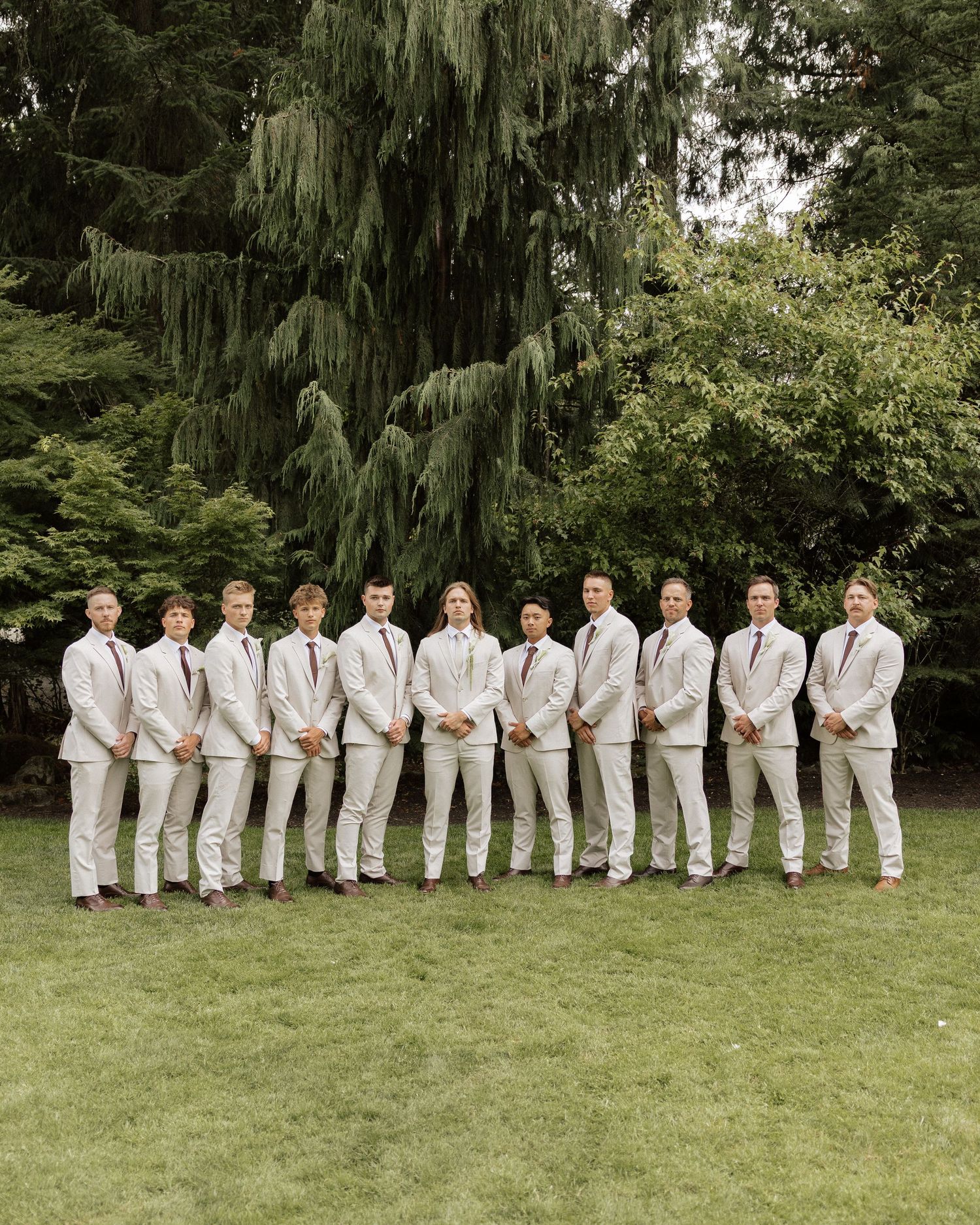 Groom and groomsmen in matching beige suits with burgundy ties posing in a row on green lawn outdoors.