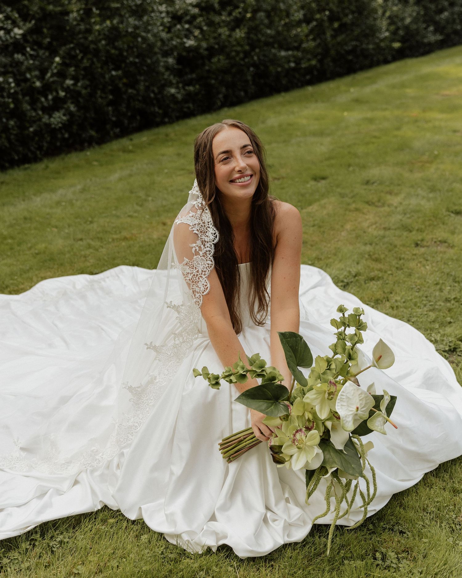 Smiling bride in strapless white gown with lace veil holding green and white bouquet while sitting on grass.