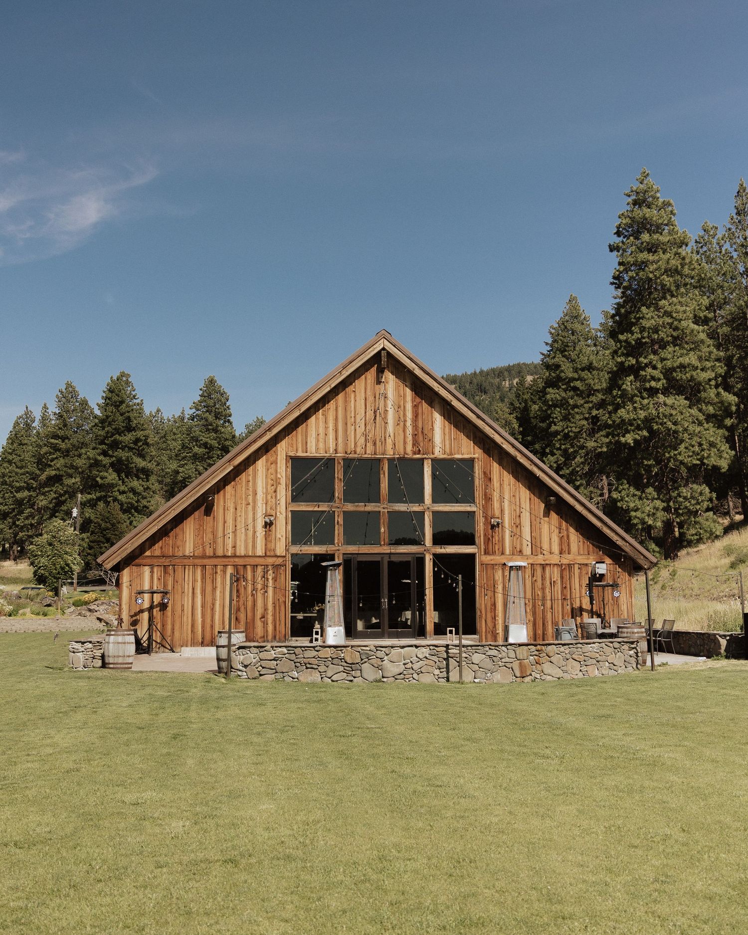 Rustic wooden barn venue with stone foundation and large windows set on a green hillside.