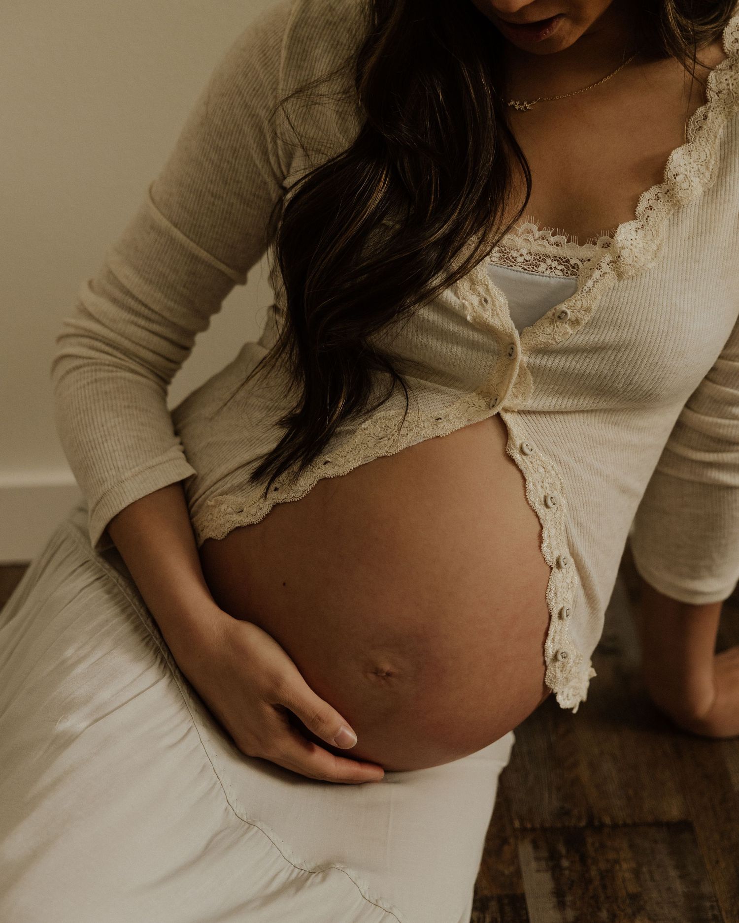 Close-up of pregnant belly with hands resting on it, wearing cream lace-trimmed top.