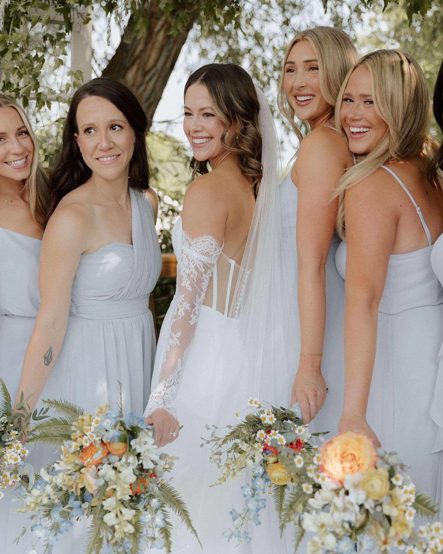 Bride in white lace dress poses with bridesmaids wearing pale blue dresses and holding floral bouquets.