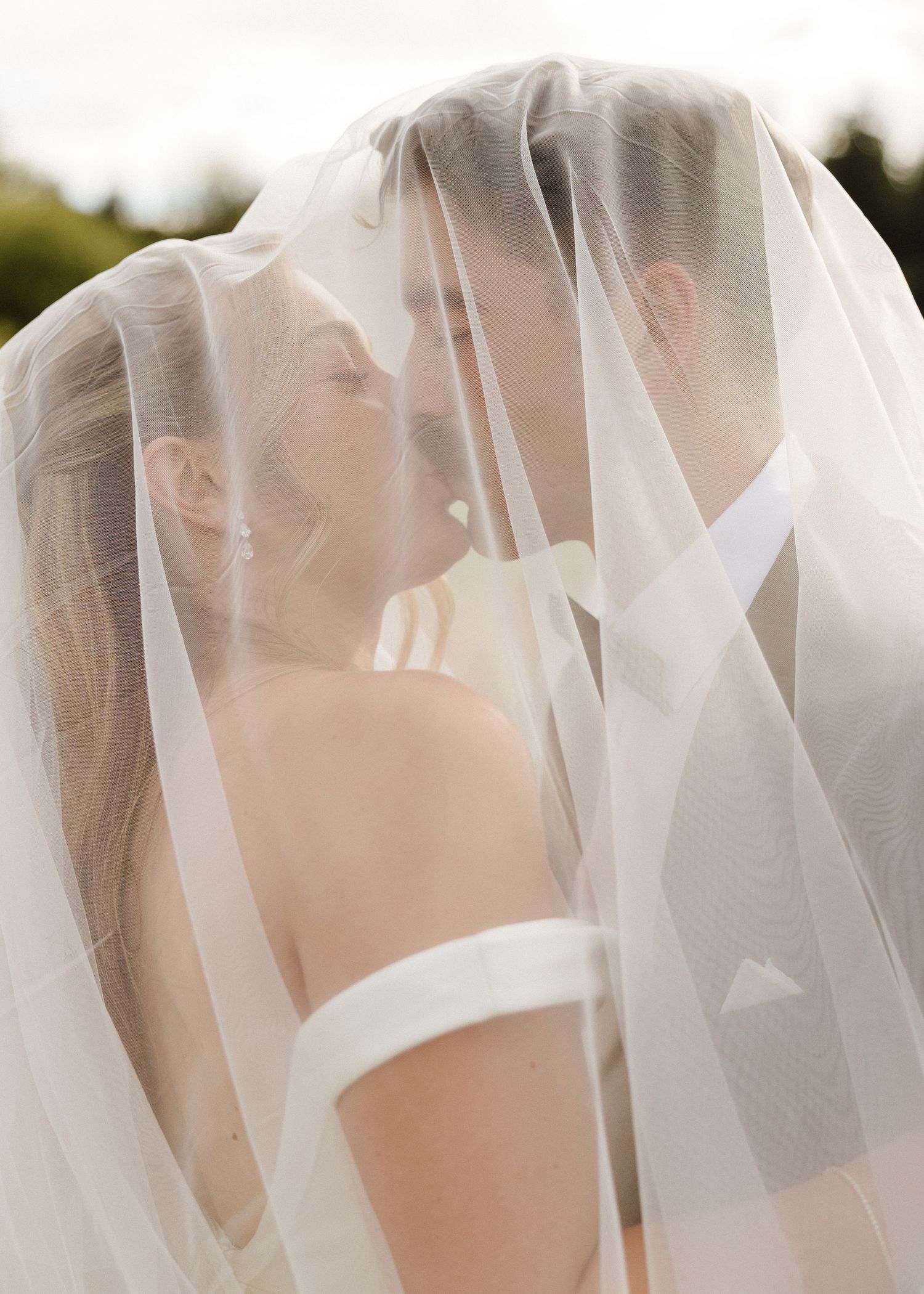 A bride and groom share an intimate moment under a flowing white wedding veil, nearly kissing in soft natural light.