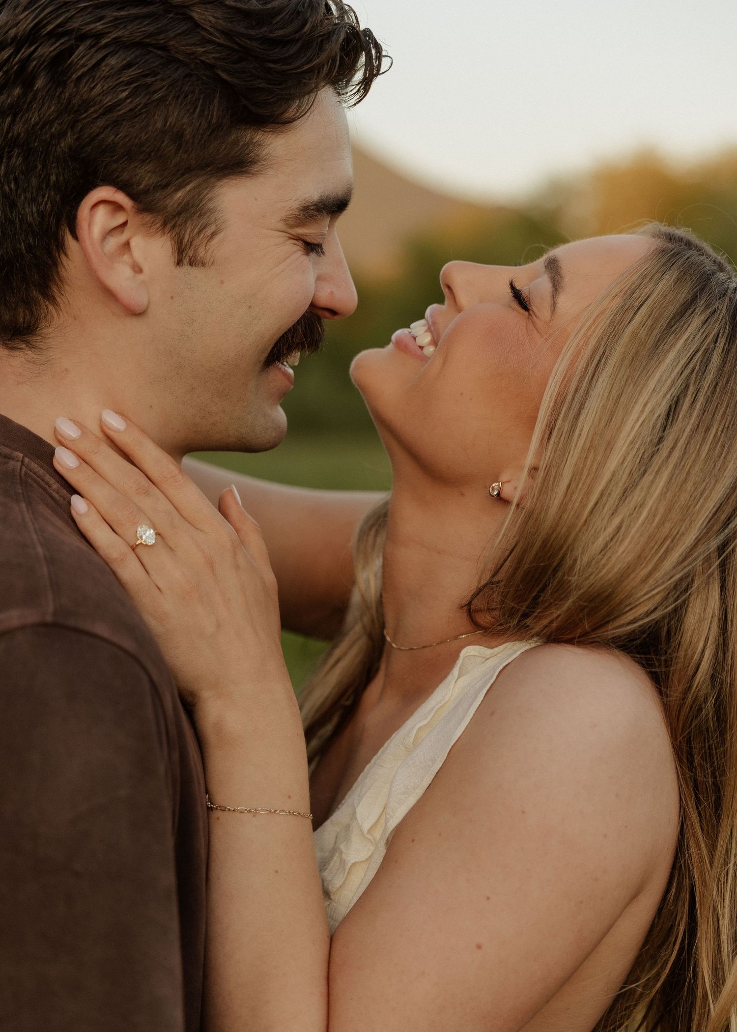 Couple sharing a joyful moment, woman laughing with engagement ring visible, golden hour outdoor setting.
