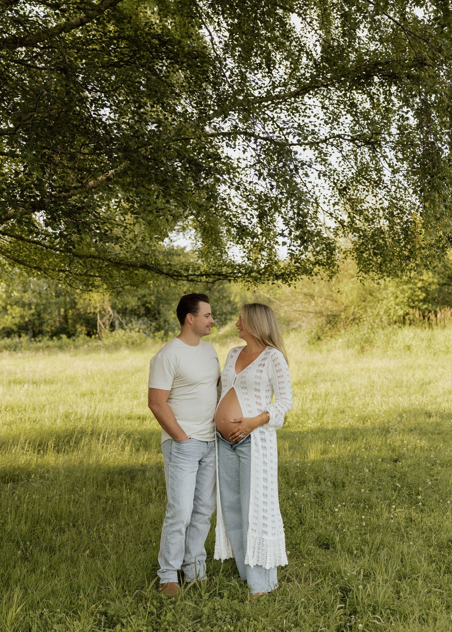 Couple sharing a tender moment under a tree in a green field, woman in white lace cardigan showing baby bump.