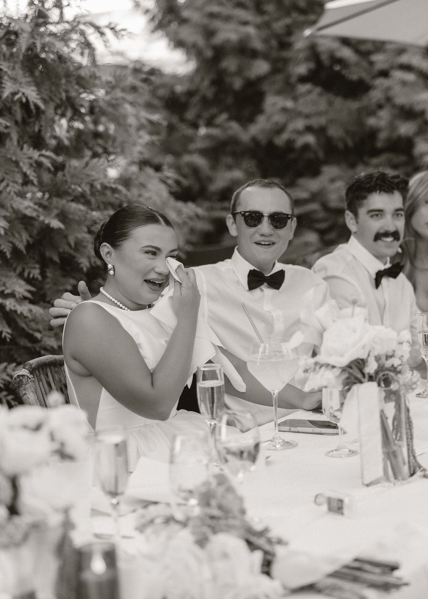 Black and white photo of bride and groom laughing at the sweetheart table during outdoor wedding reception.