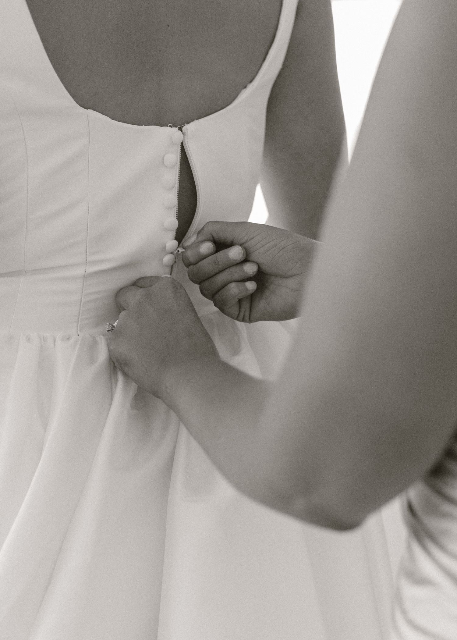 Black and white photo of a bridesmaid buttoning the back of a bride's white wedding dress.