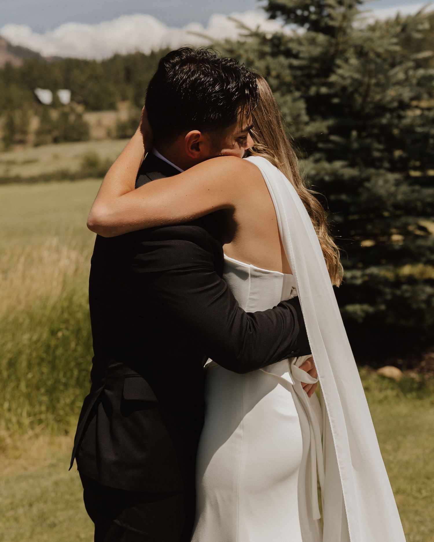 Close embrace between newlyweds in a rustic field with evergreen trees in the background against a cloudy sky.
