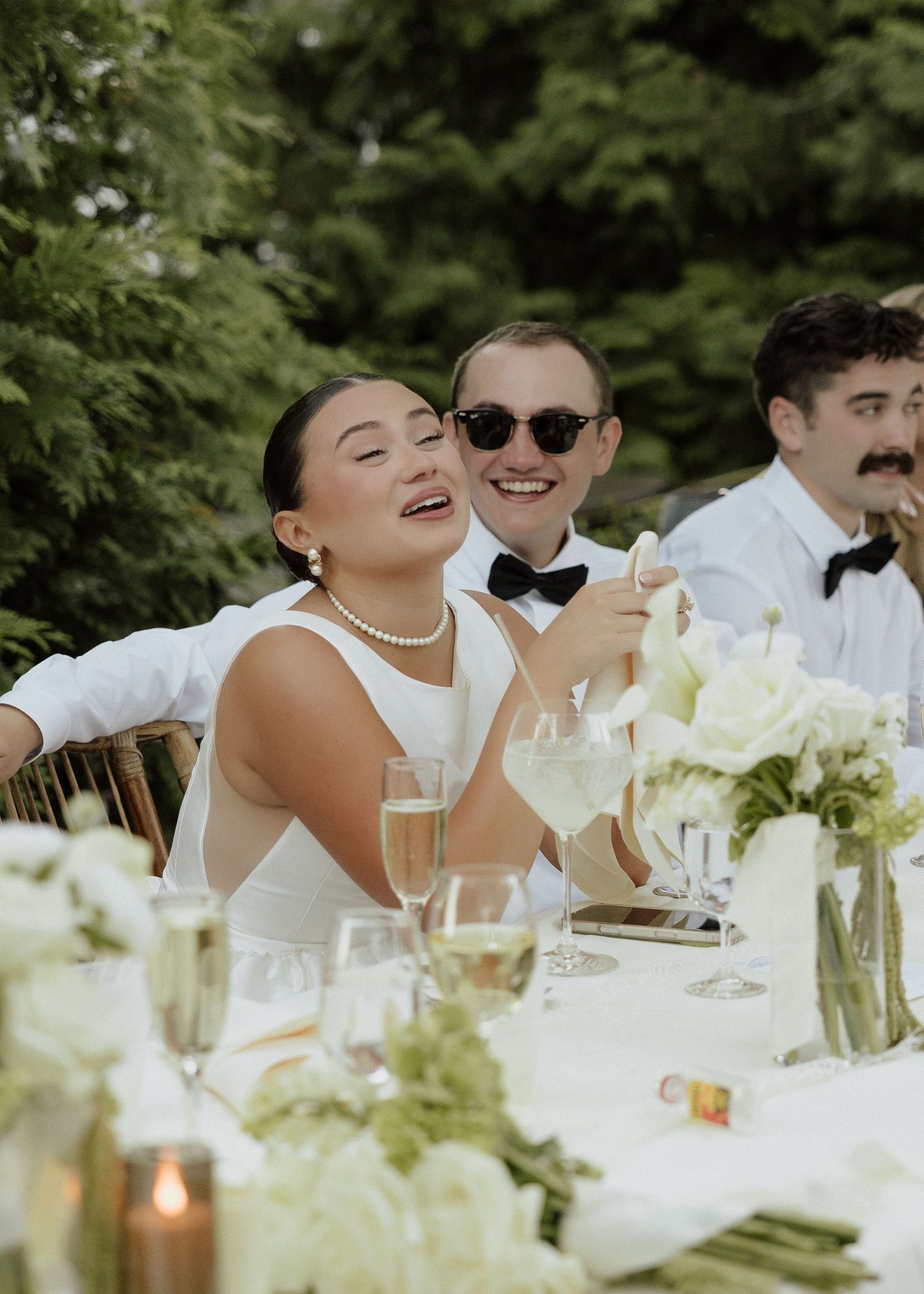Bride laughing at outdoor wedding reception table with champagne, white floral centerpieces, and guests in bow ties.