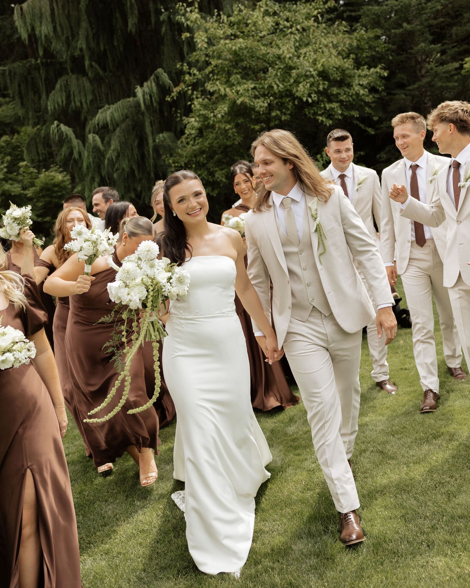 Bride in white strapless gown and groom in beige suit walk with wedding party in brown dresses outdoors.