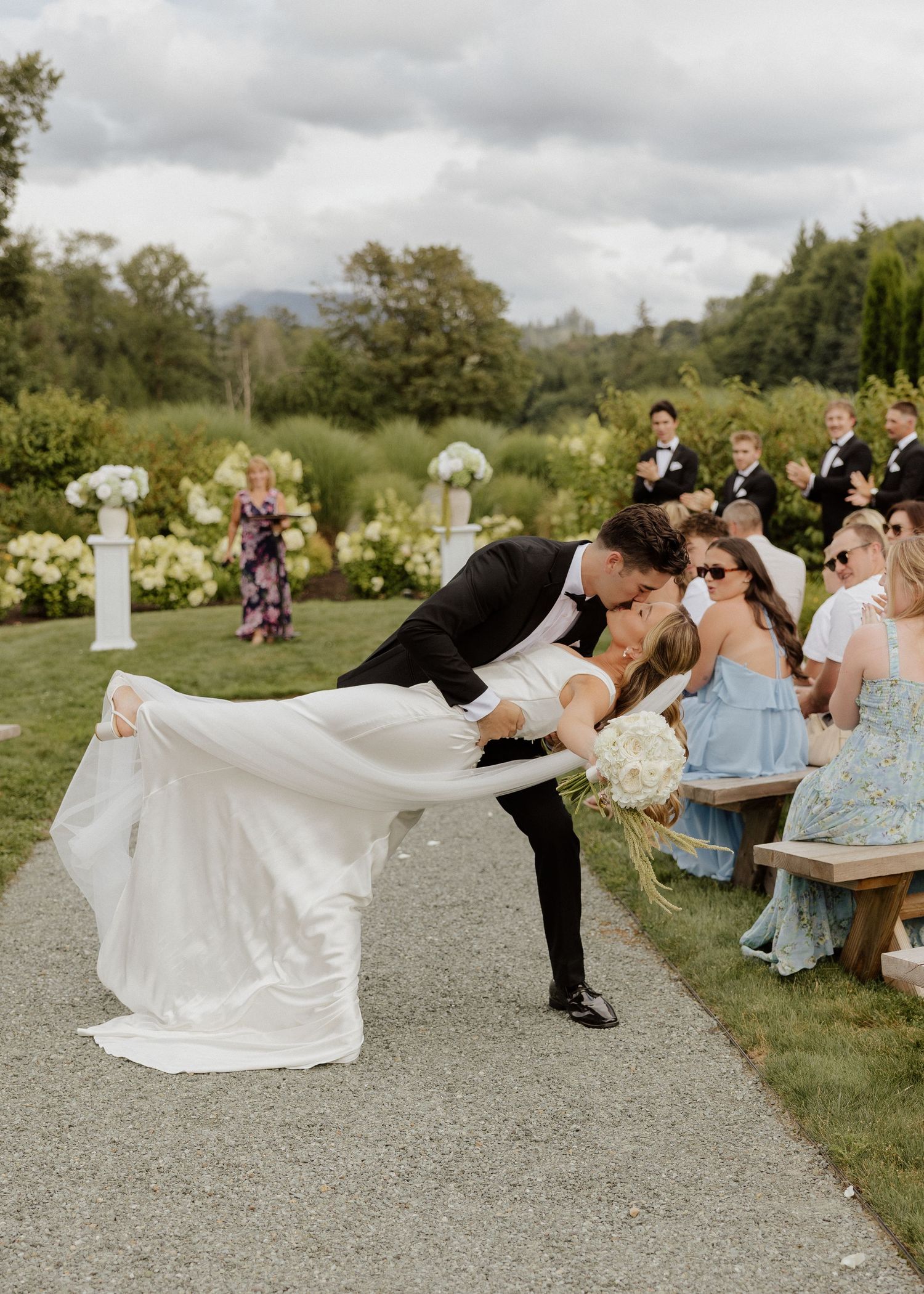 Groom dips bride for a kiss while guests cheer during outdoor wedding ceremony recessional.