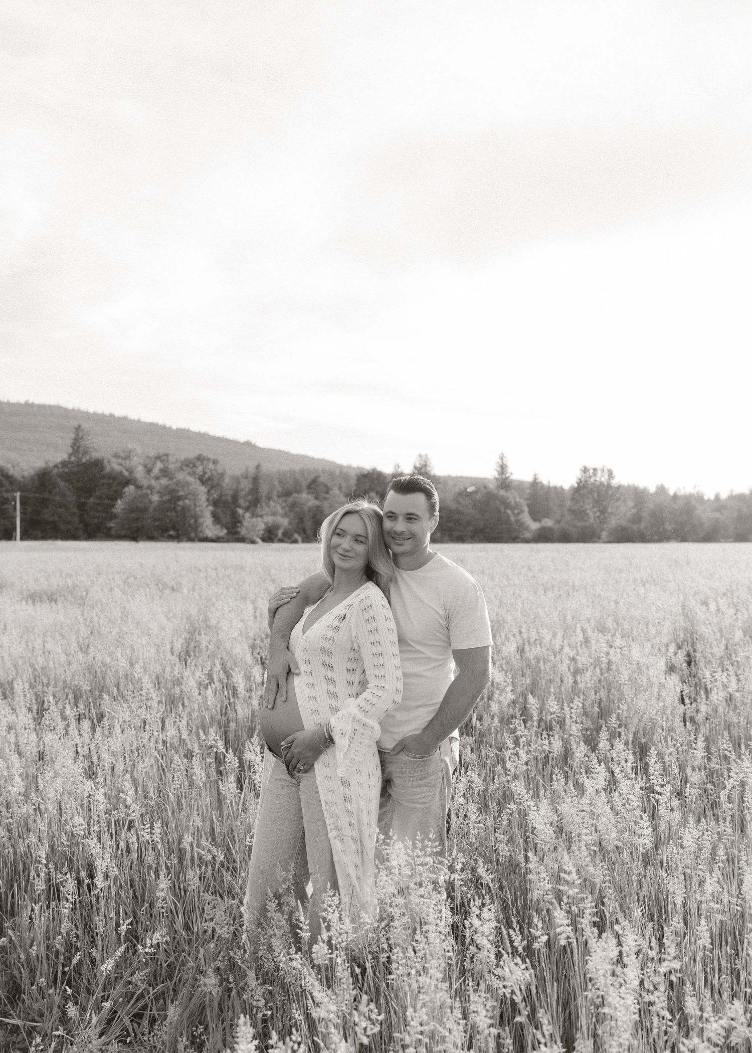 Couple embracing in a field during a black and white maternity photo shoot with scenic hills.