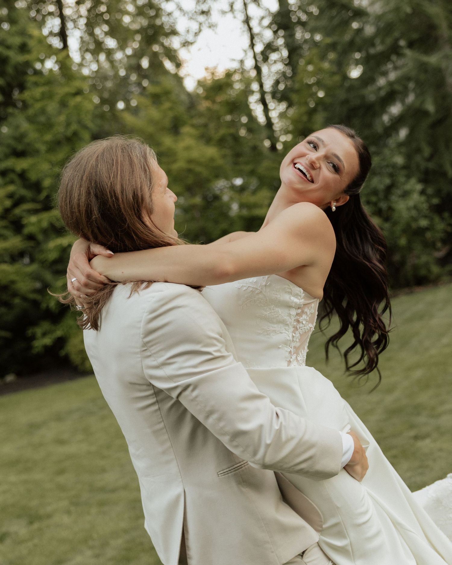 A joyful bride in a white gown laughs as her partner in a cream suit lifts her outdoors with lush green trees behind.