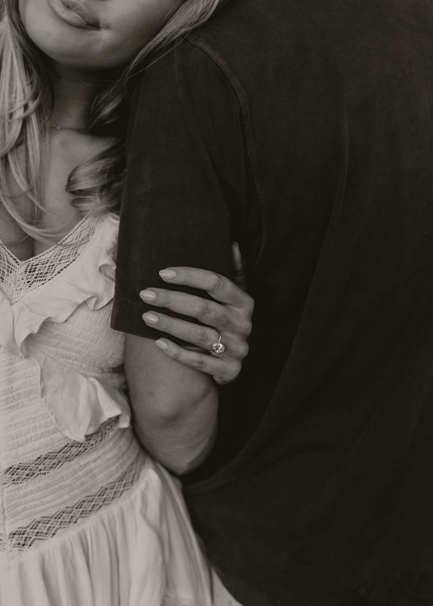 Close-up of a woman in a white lace dress holding a mans arm, showcasing an engagement ring in black and white.