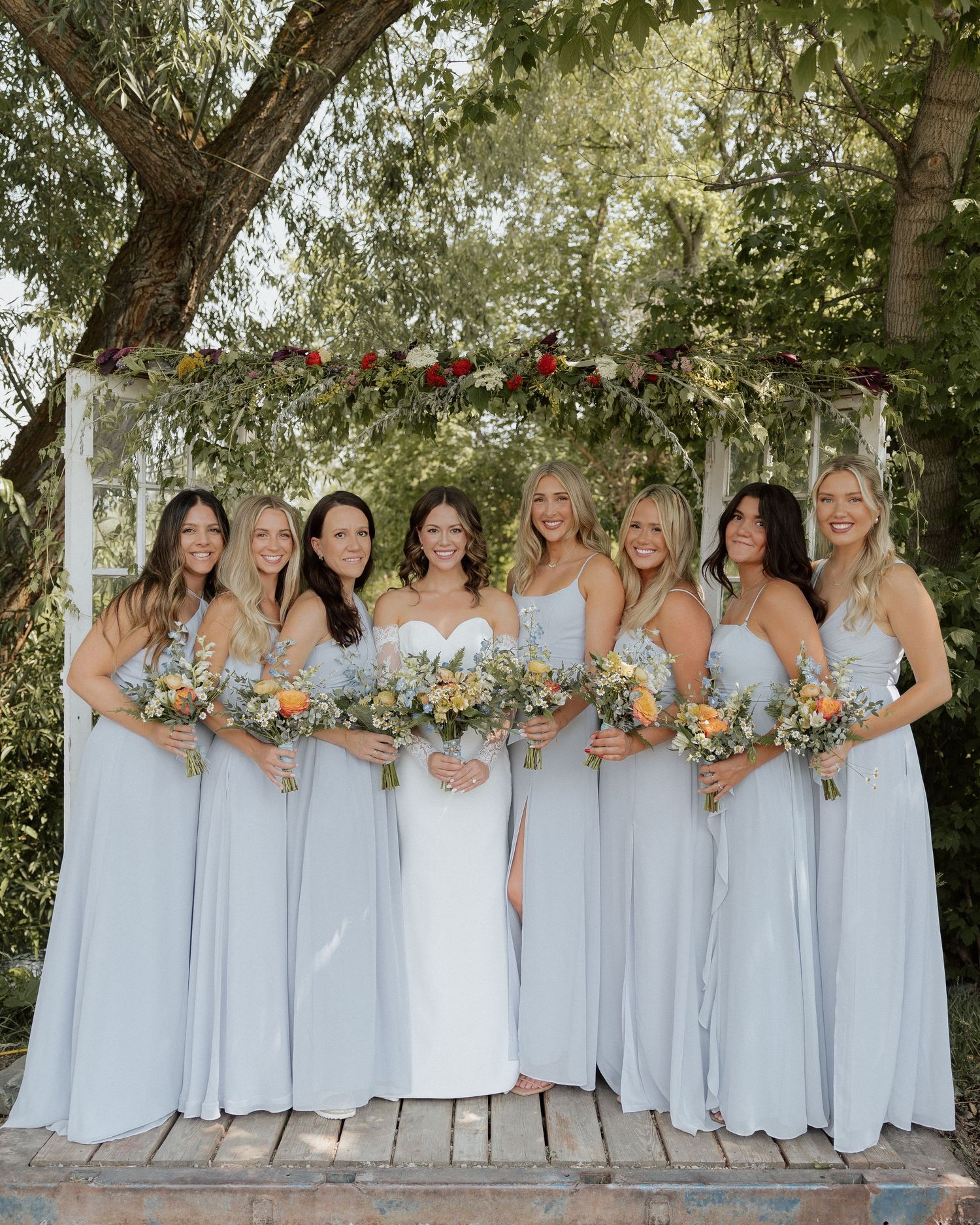 Bridal party in light blue dresses standing with bride under floral arch.