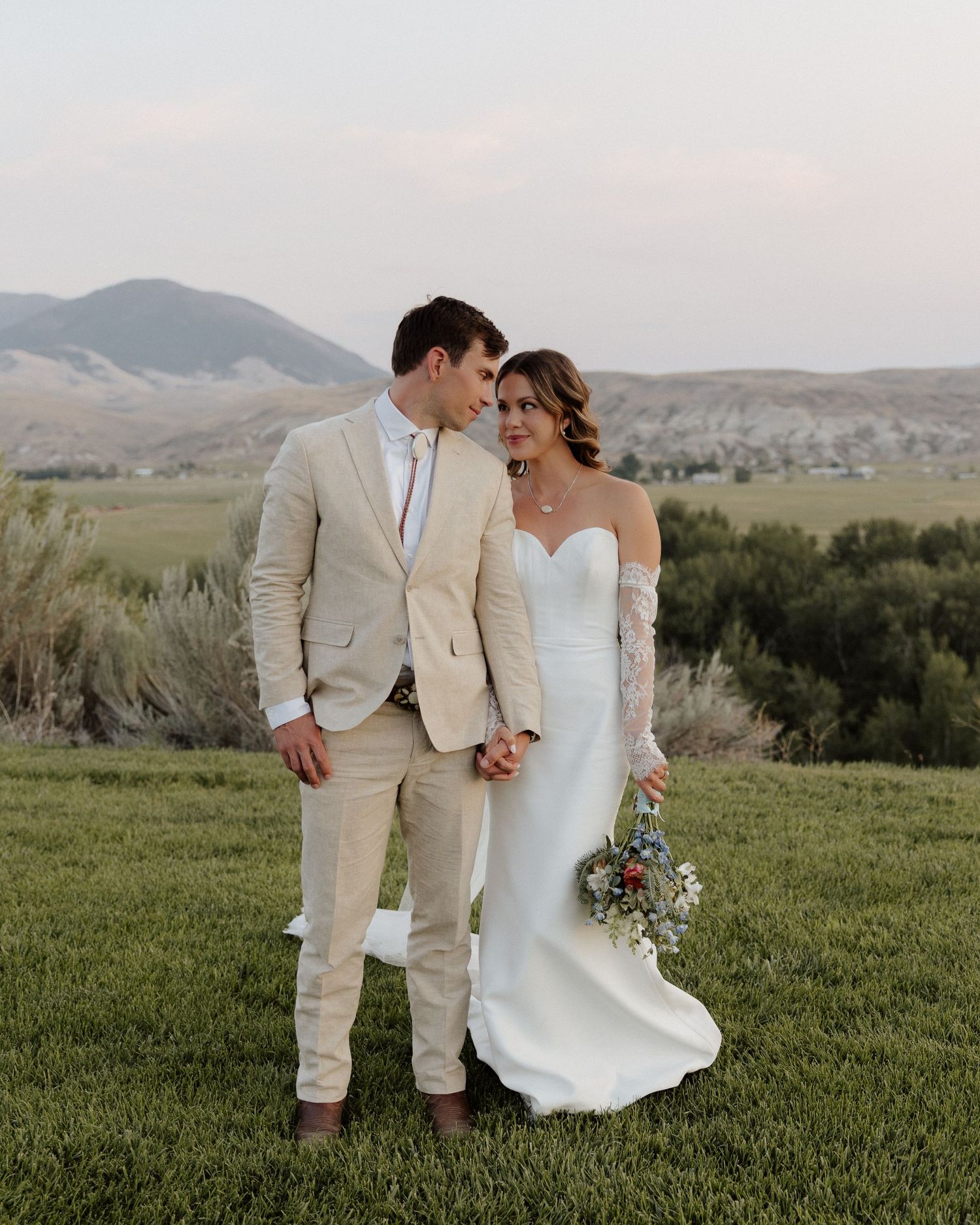 Bride and groom share tender moment on grassy hillside in matching beige and white attire.