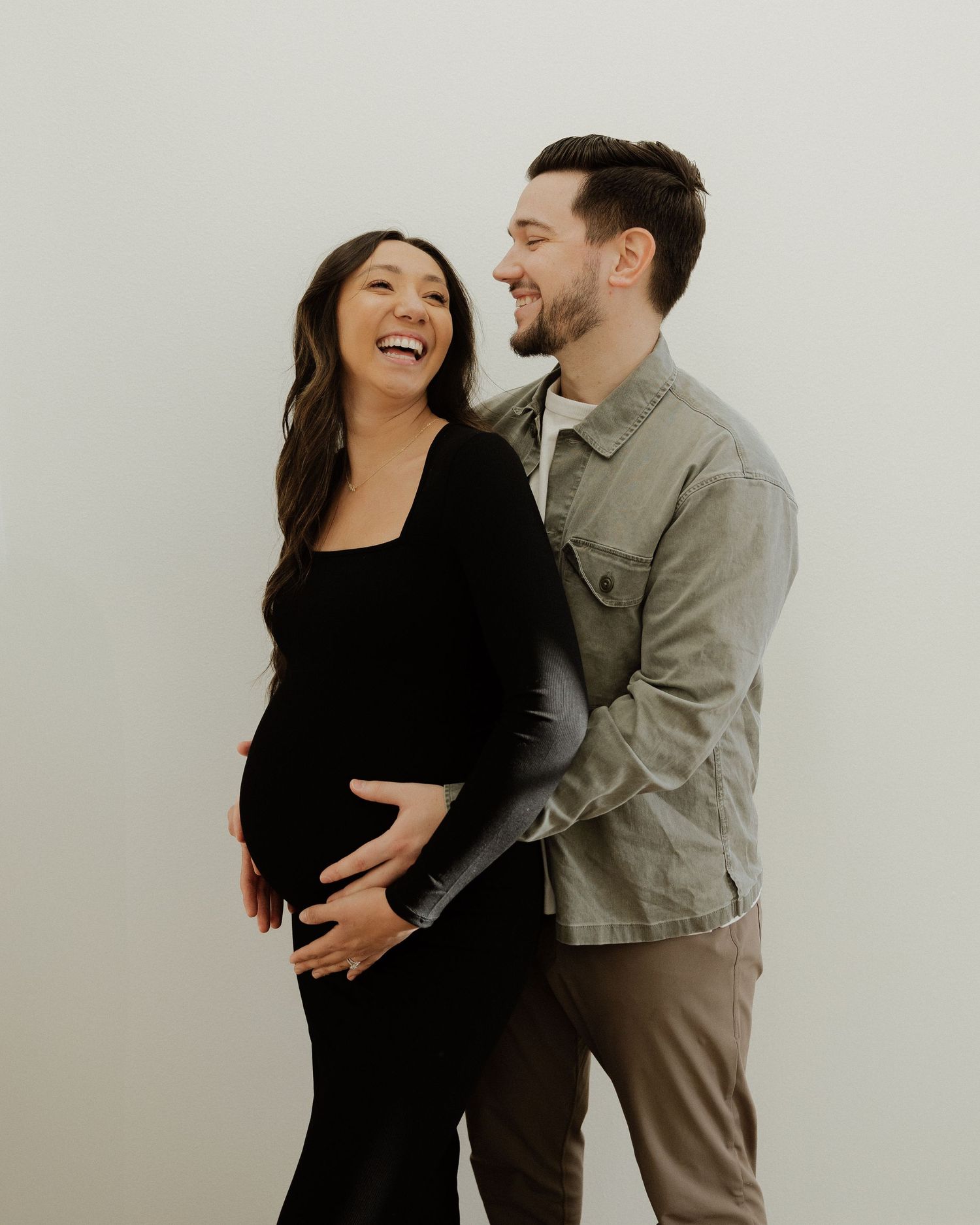 Joyful couple sharing a candid moment during maternity photoshoot against white wall.