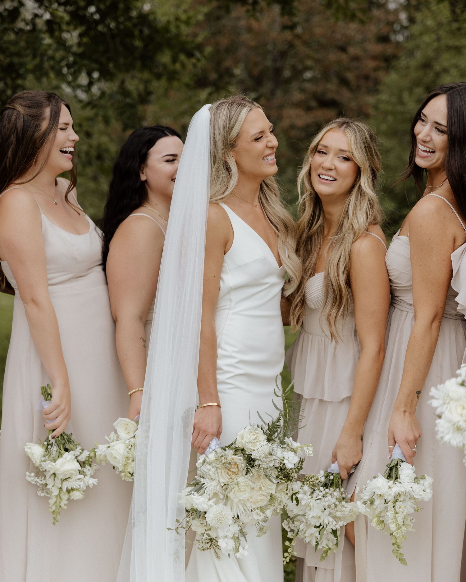 Wedding party members in neutral dresses share genuine laughter while holding white bouquets.
