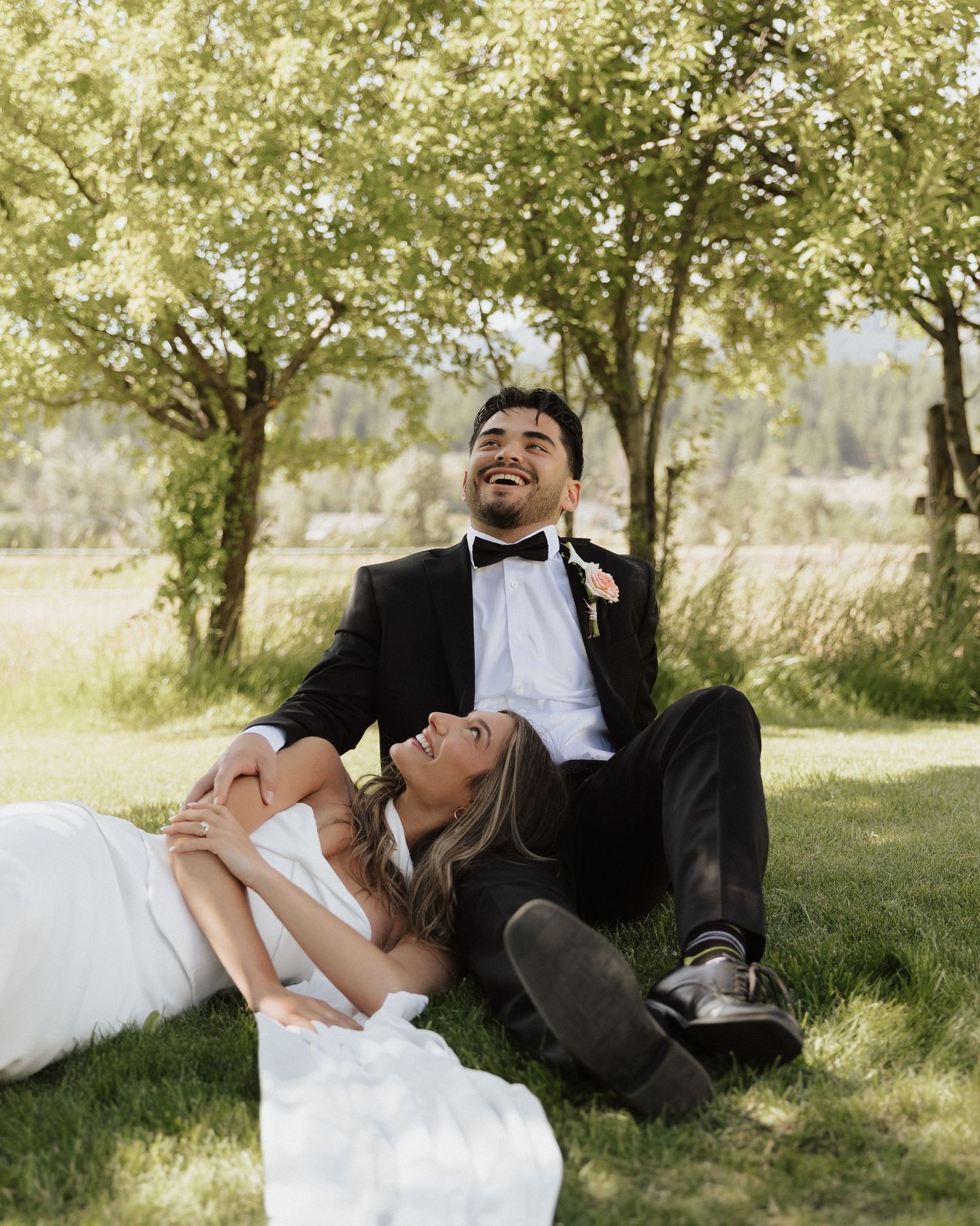 A romantic wedding photoshoot on lush grass beneath trees showing playful poses in formal attire.