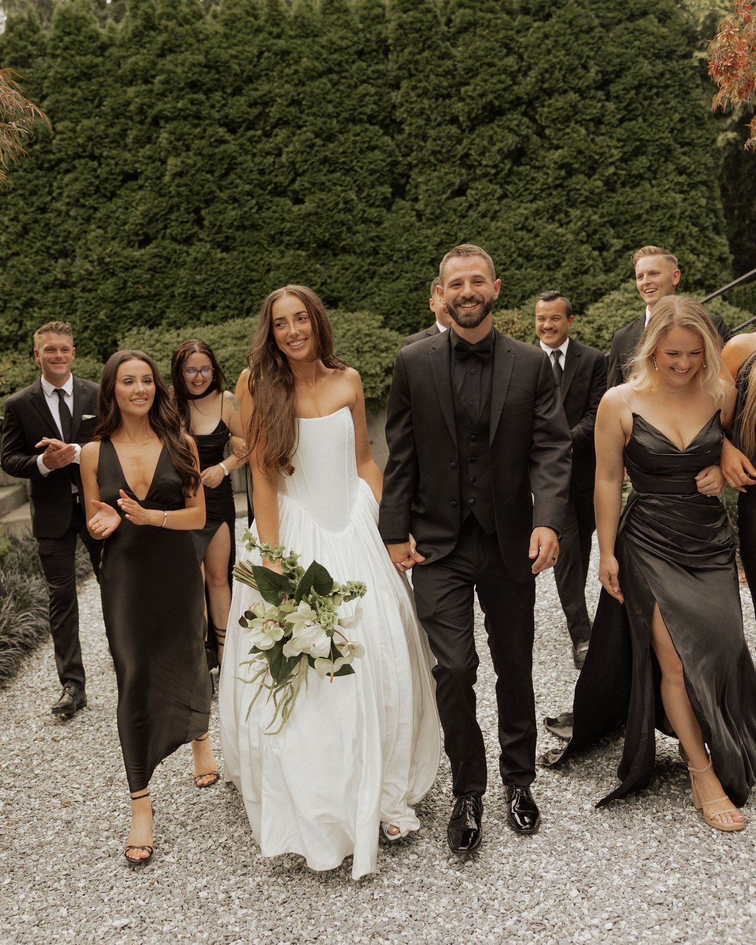 Bride and groom walk with wedding party in black attire, bride holding white floral bouquet on gravel path.