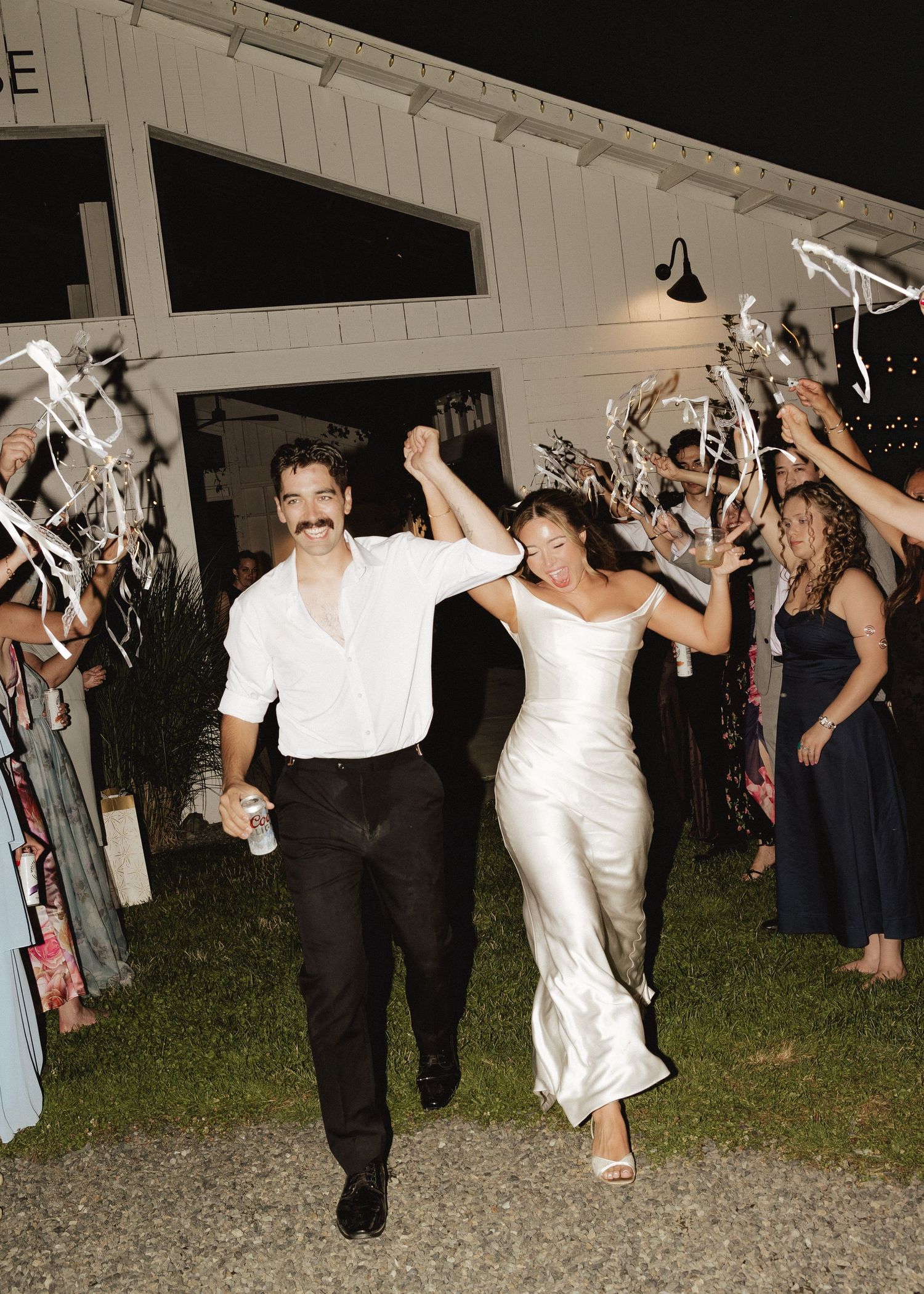 Bride and groom exit wedding venue through cheering guests waving ribbon wands under night sky celebration.