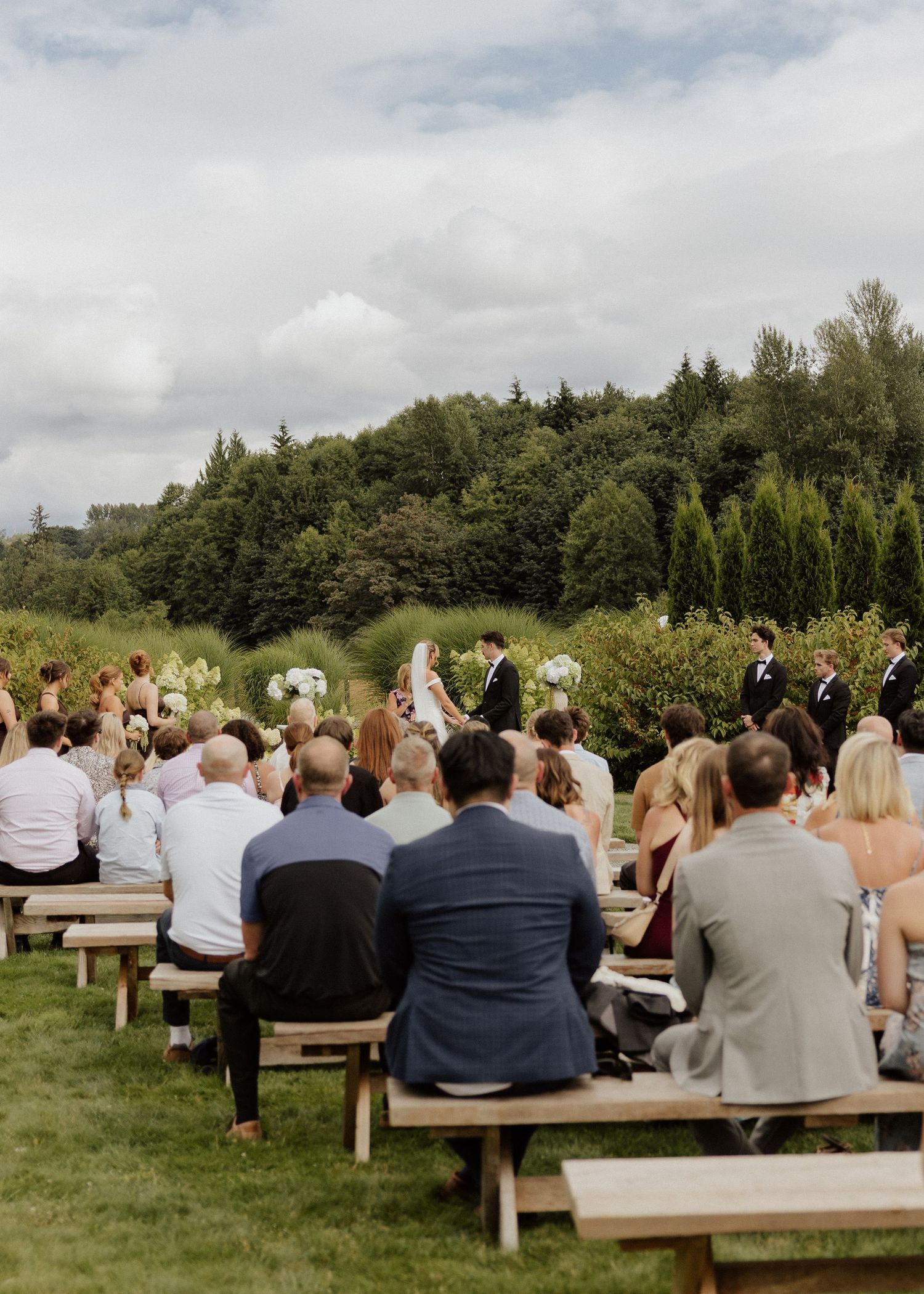 Outdoor wedding ceremony with guests seated on wooden benches facing couple at altar surrounded by lush green trees.