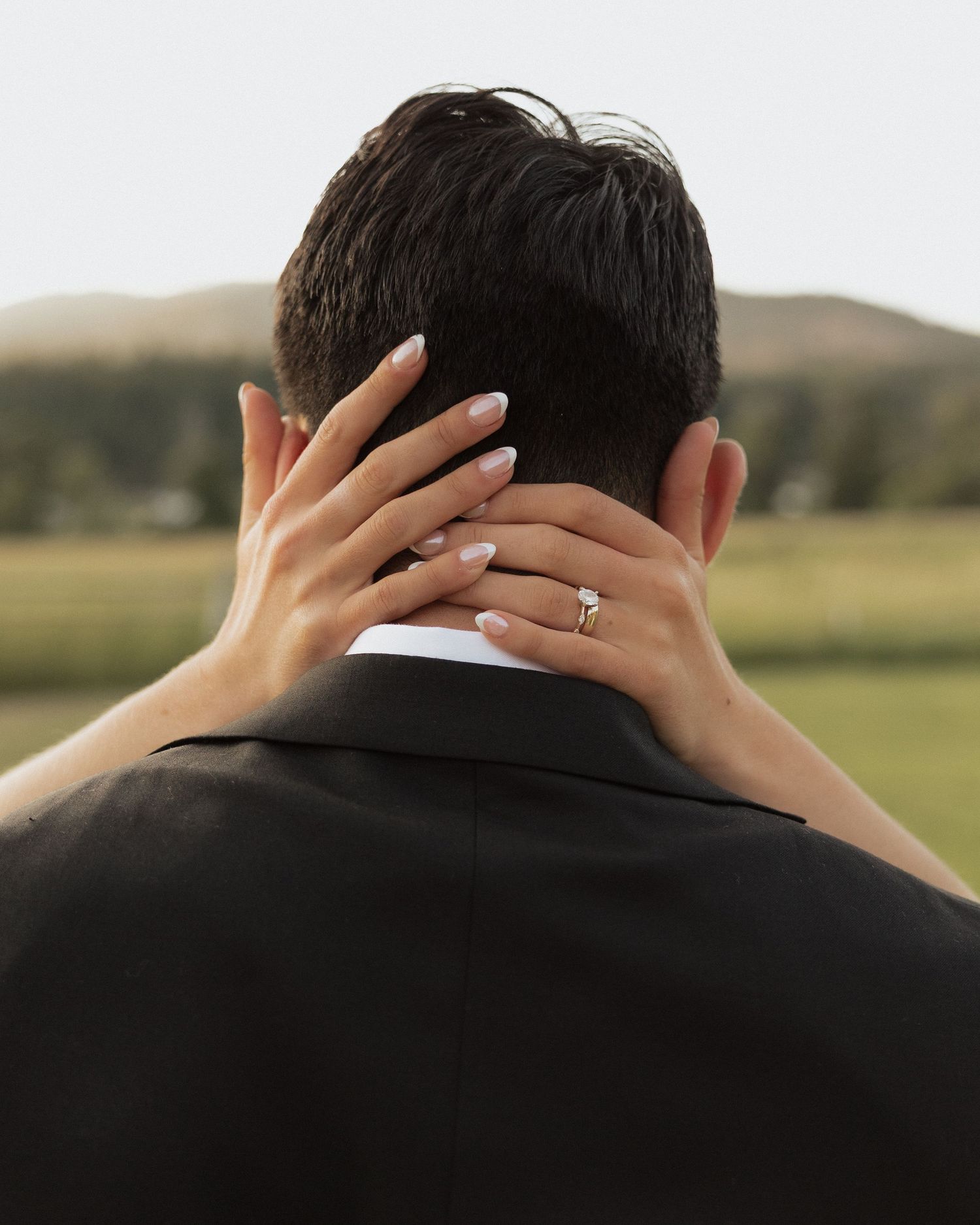 A bride's hand with an engagement ring rests on a groom's neck as they embrace outdoors with green hills behind.