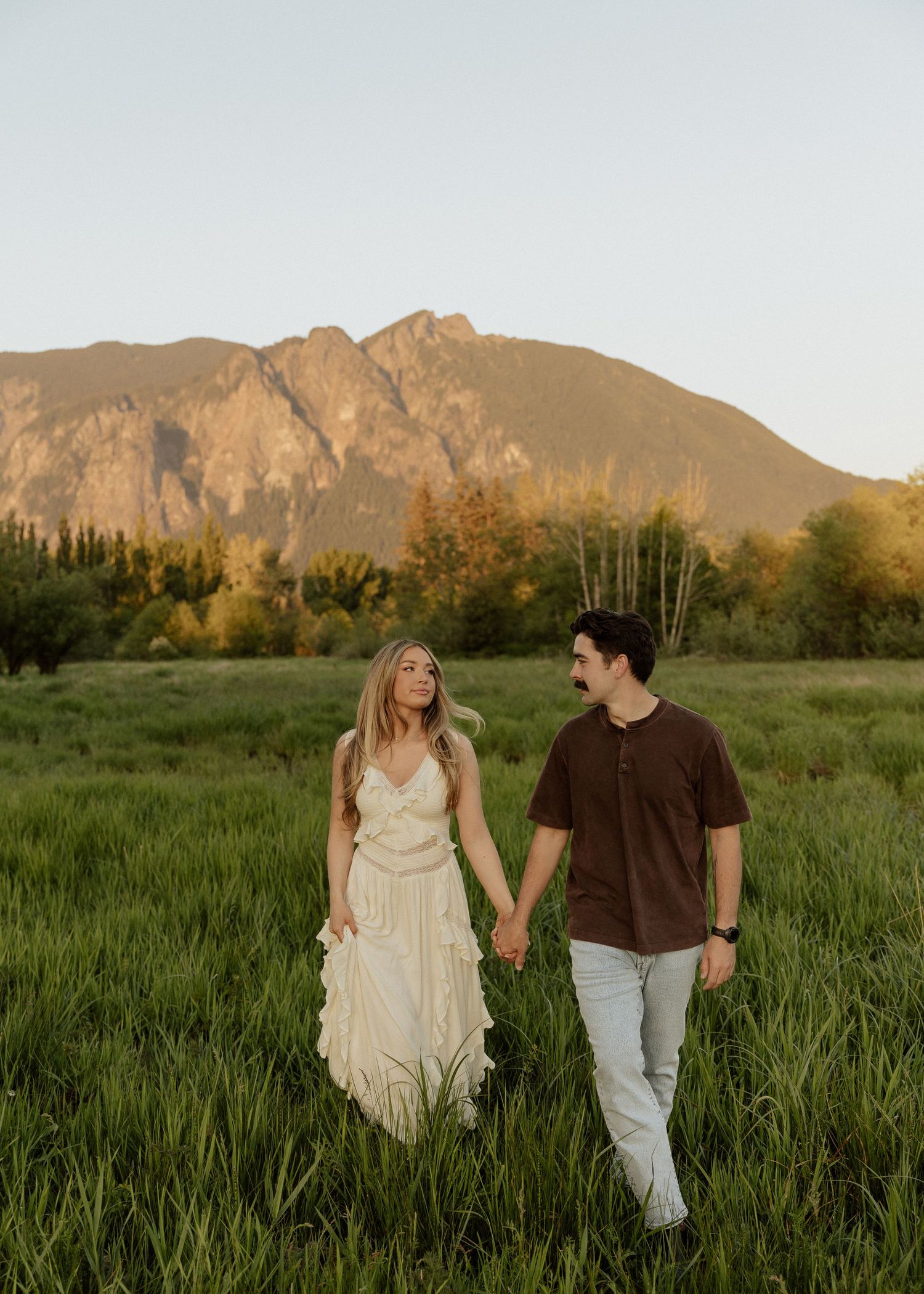 A couple holds hands walking through a green meadow with a scenic mountain backdrop at sunset.