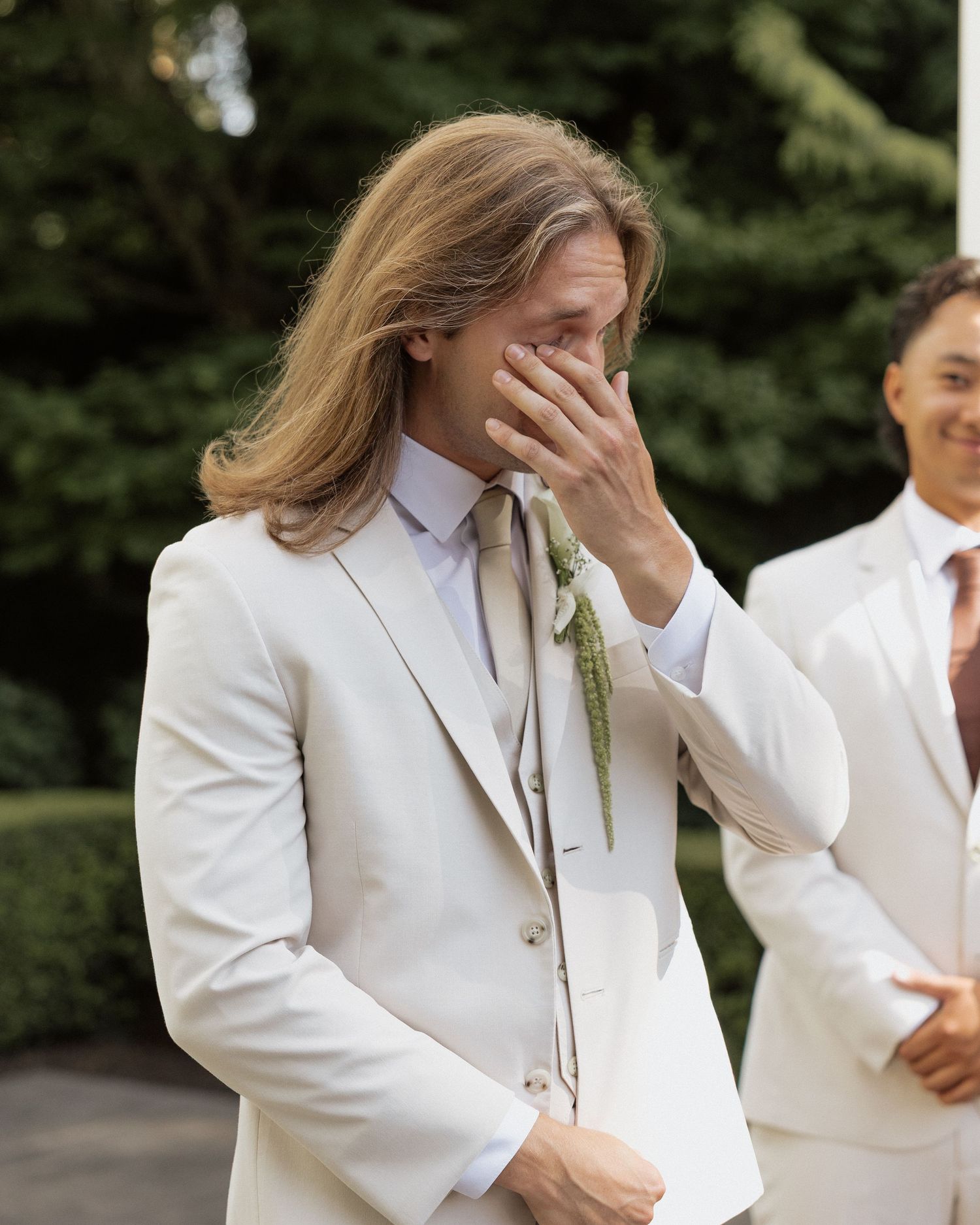 Emotional person in white suit with sage green tie covering mouth at outdoor wedding ceremony.