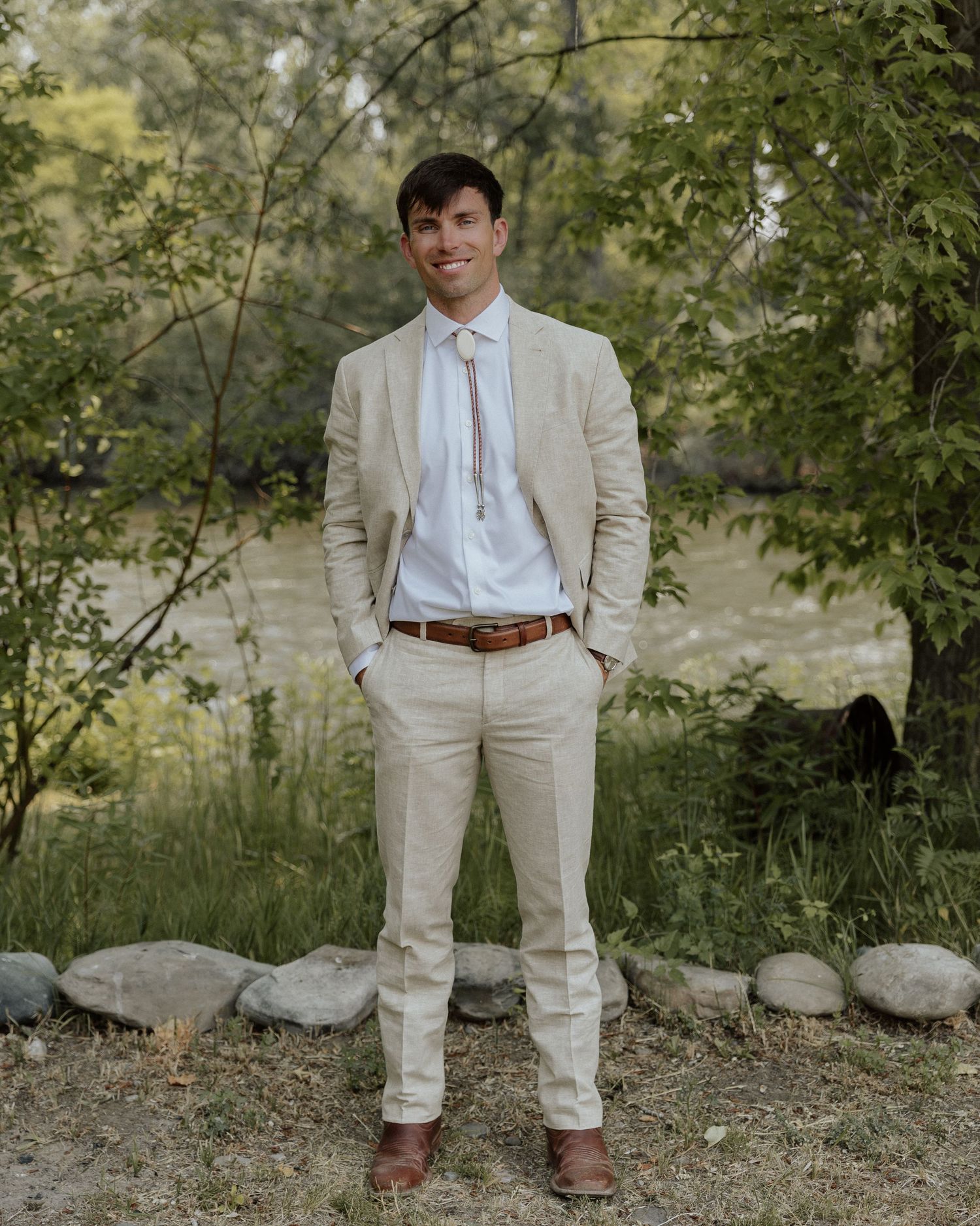 Groom in a light beige suit and tie standing outdoors in a natural setting.