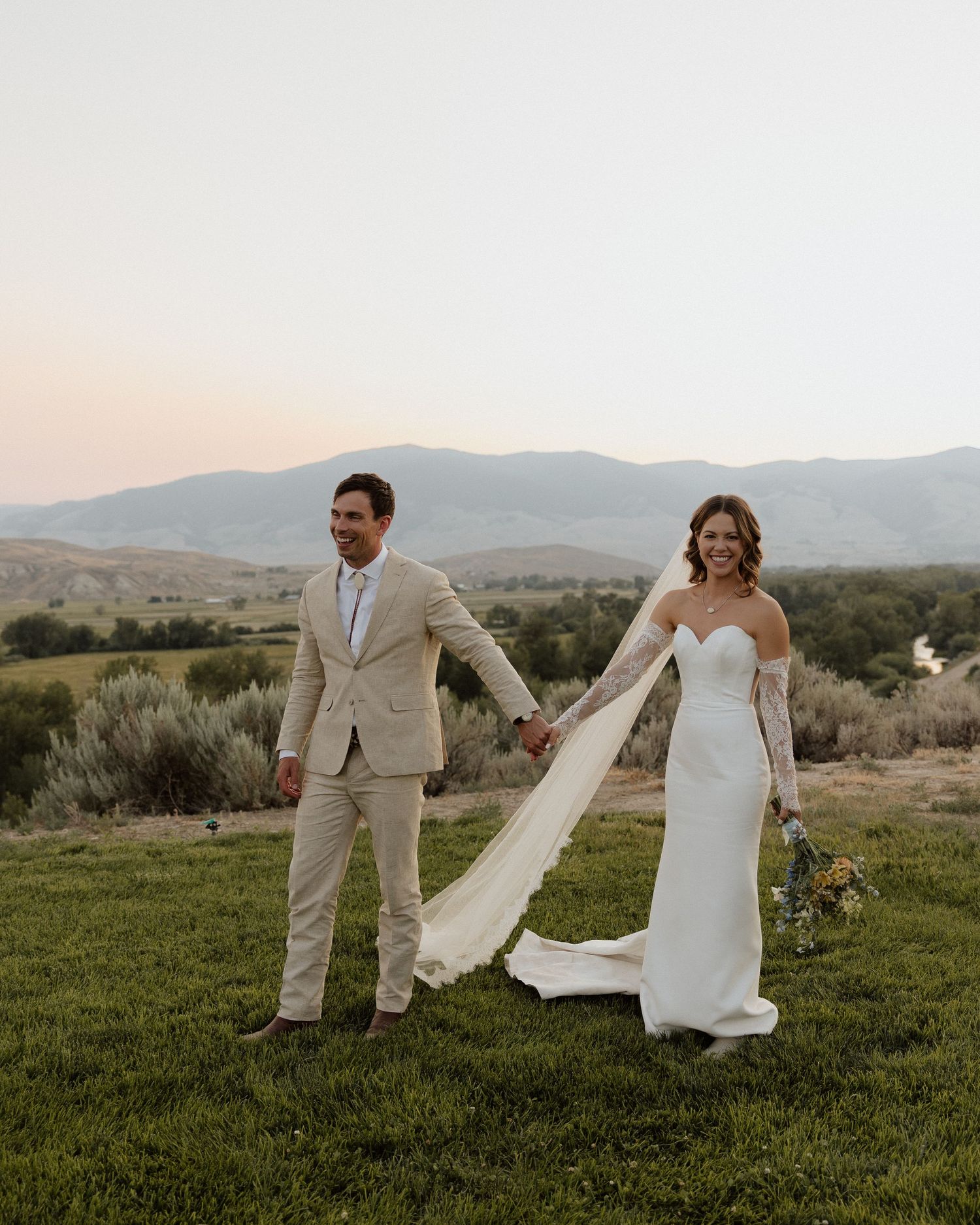 Couple in wedding attire pose playfully on grassy hilltop with flowing veil.