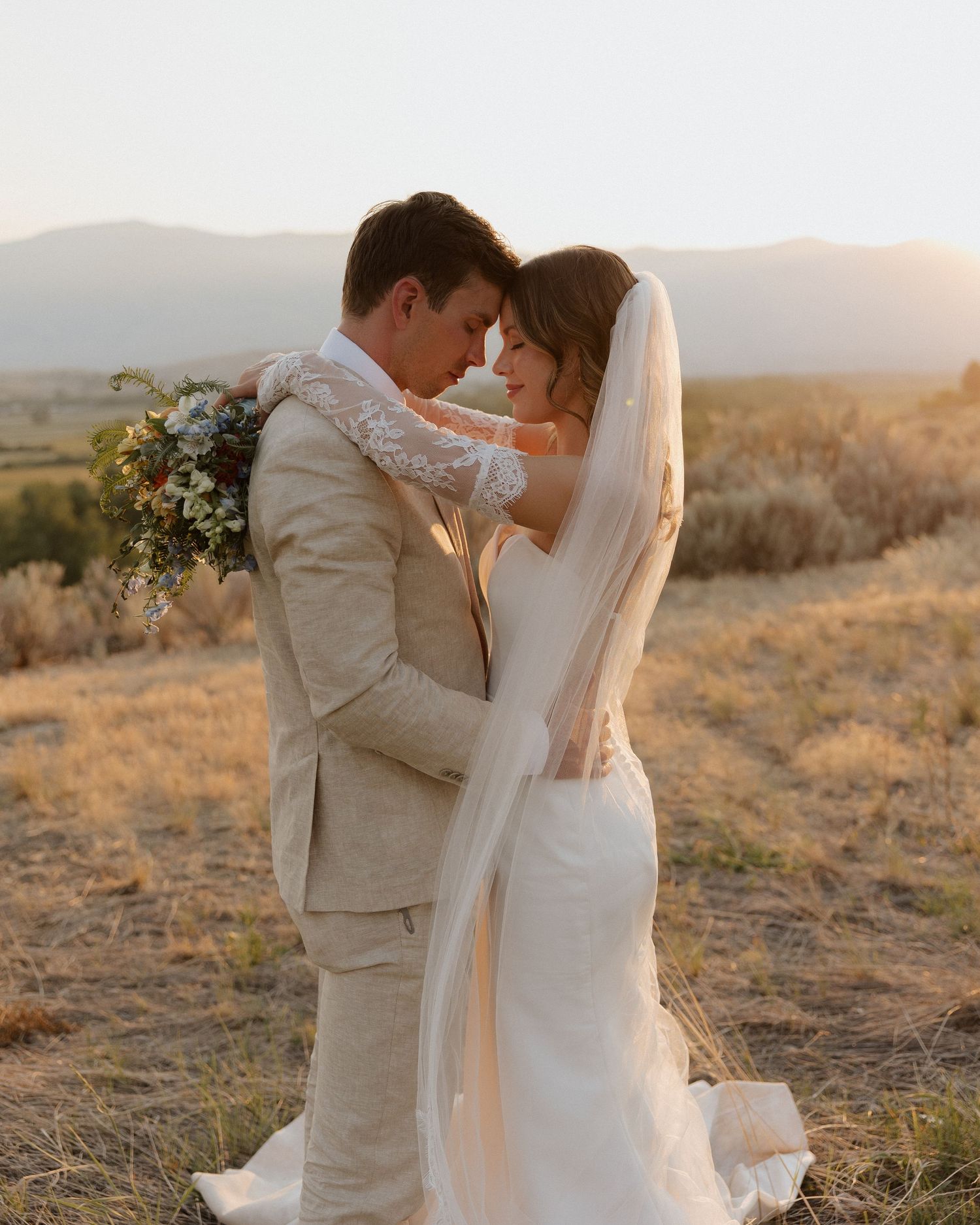 Couple in light-colored wedding attire embrace against mountainous sunset backdrop.