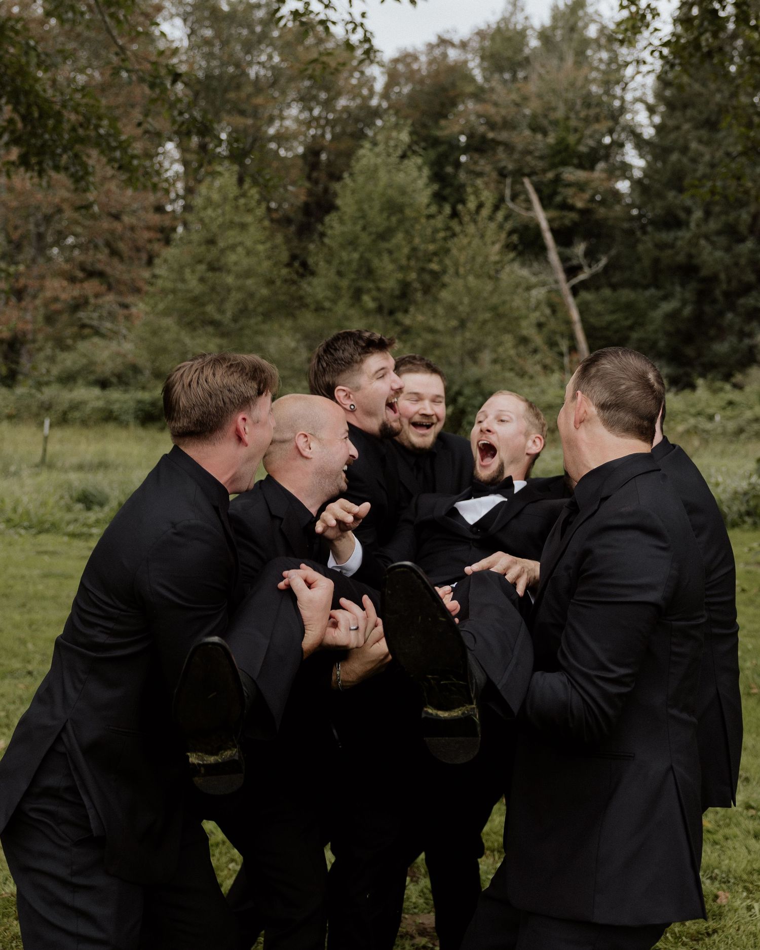 Groomsmen in black tuxedos huddle together laughing joyfully during outdoor wedding photos.