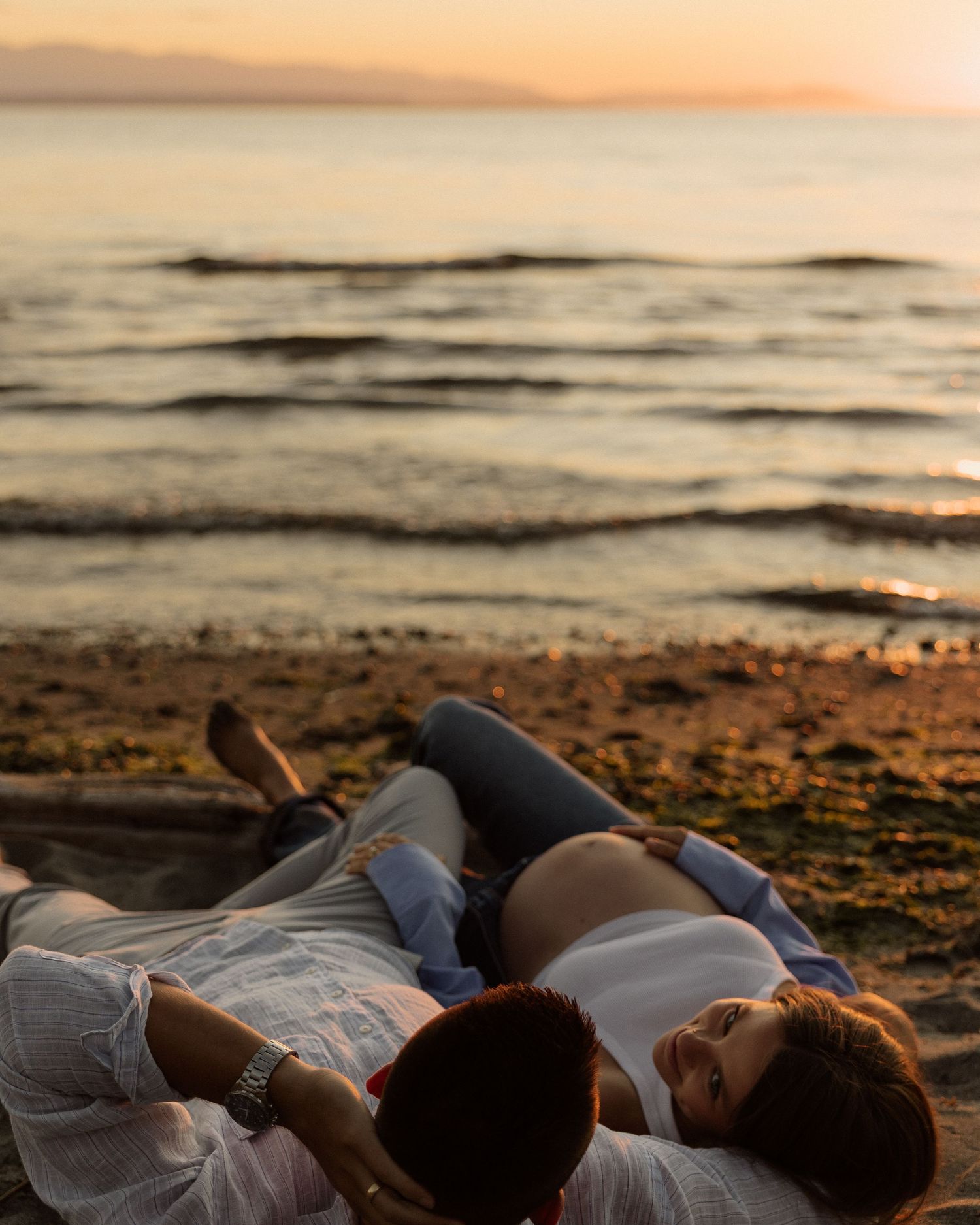 Expectant couple relaxing on beach during golden hour maternity photoshoot.