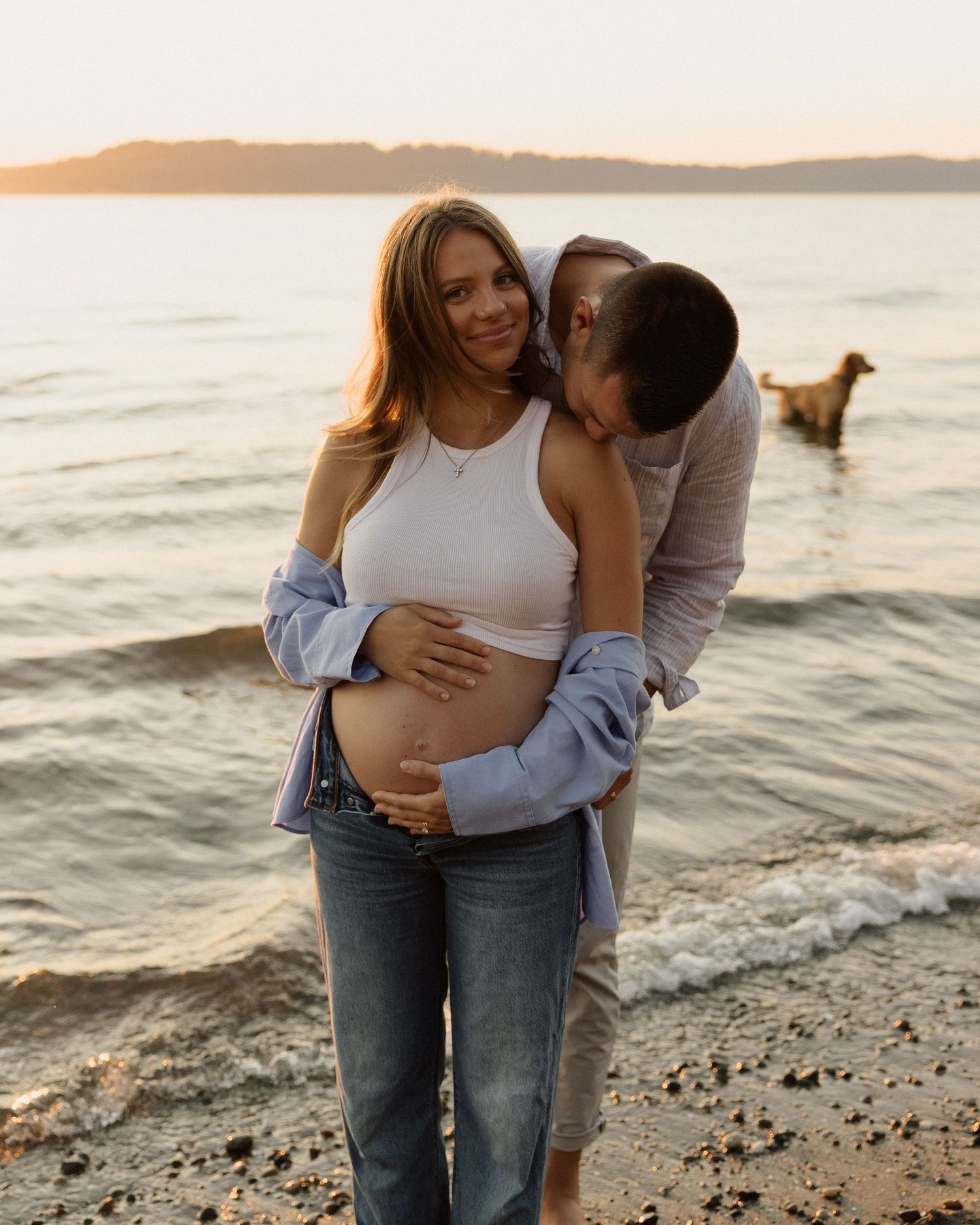 A couple sharing a tender moment during their beach maternity photoshoot with a dog in the background.