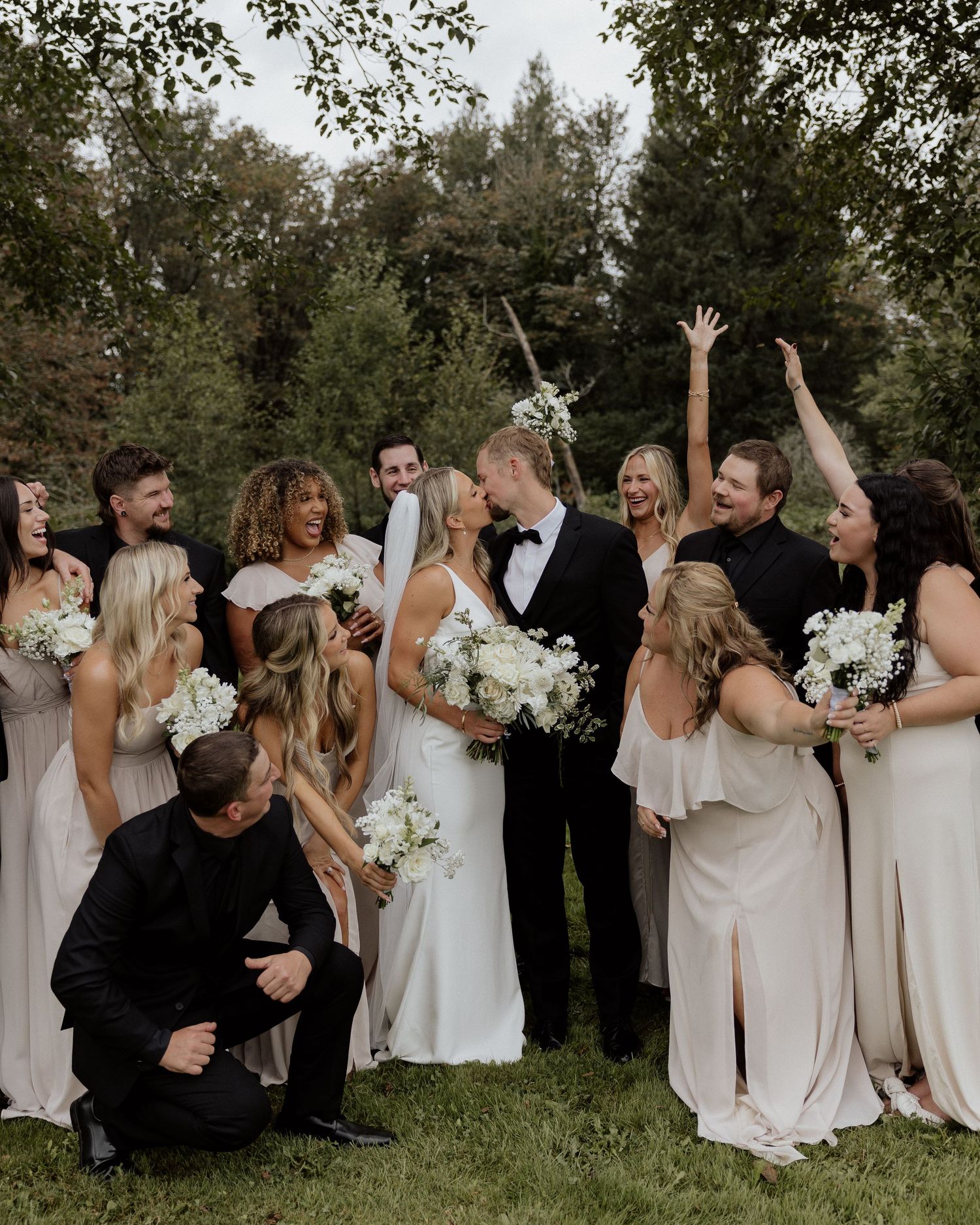 Wedding party in light neutral dresses celebrates with newlyweds in outdoor setting.