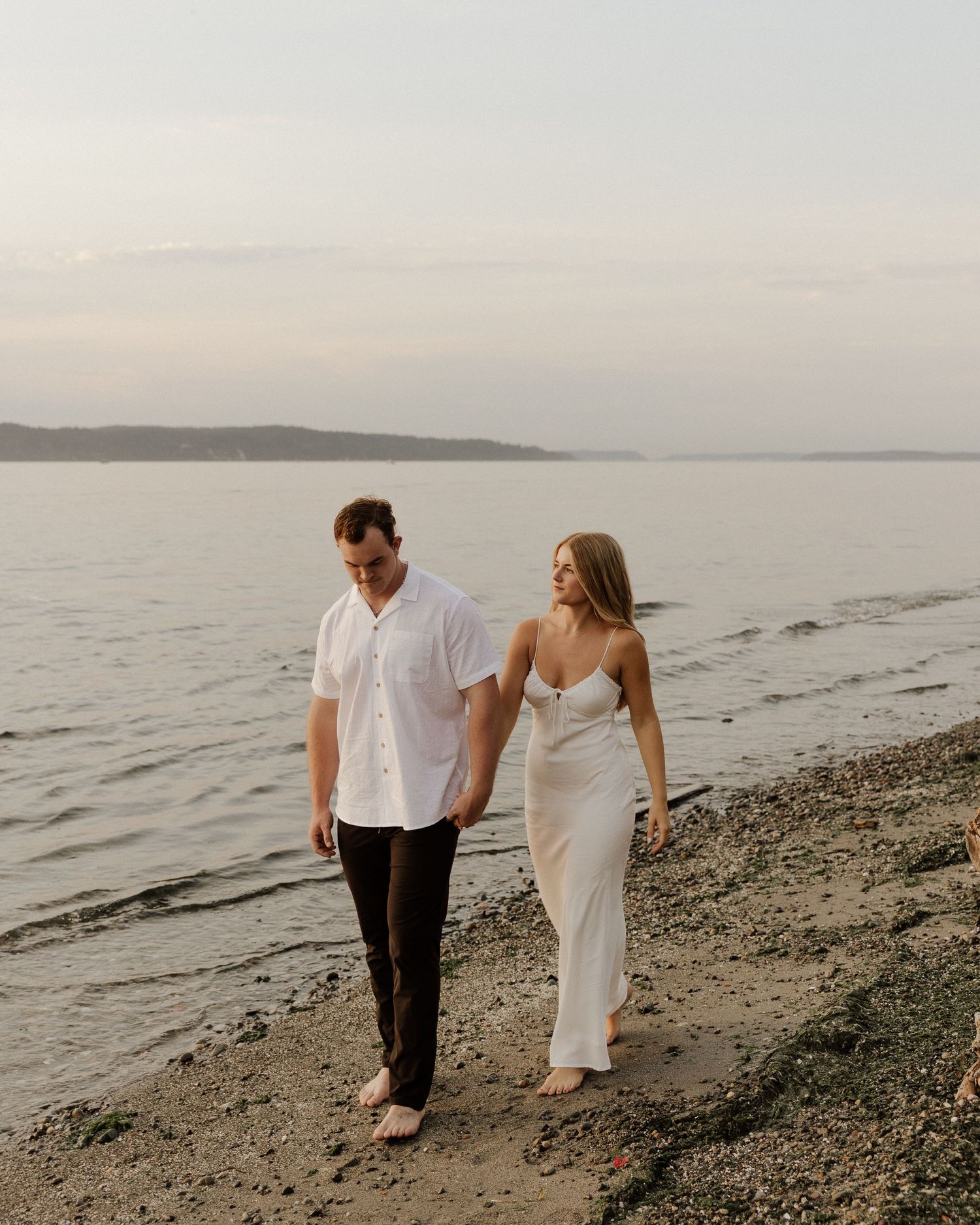 A couple strolls hand in hand along a serene beach at sunset wearing elegant white attire.