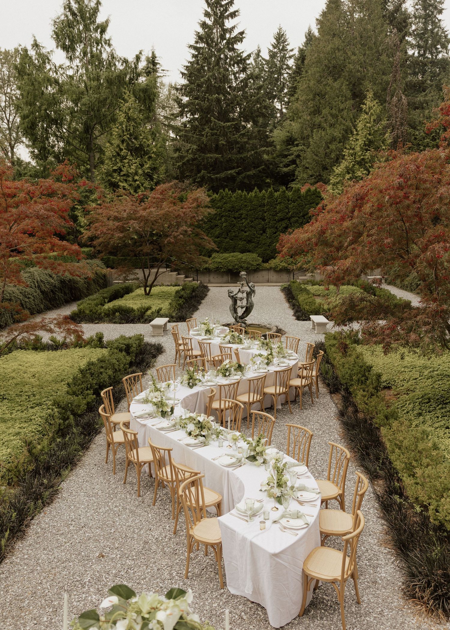Elegant outdoor wedding reception with long white tables and gold chairs in a formal garden surrounded by autumn trees.