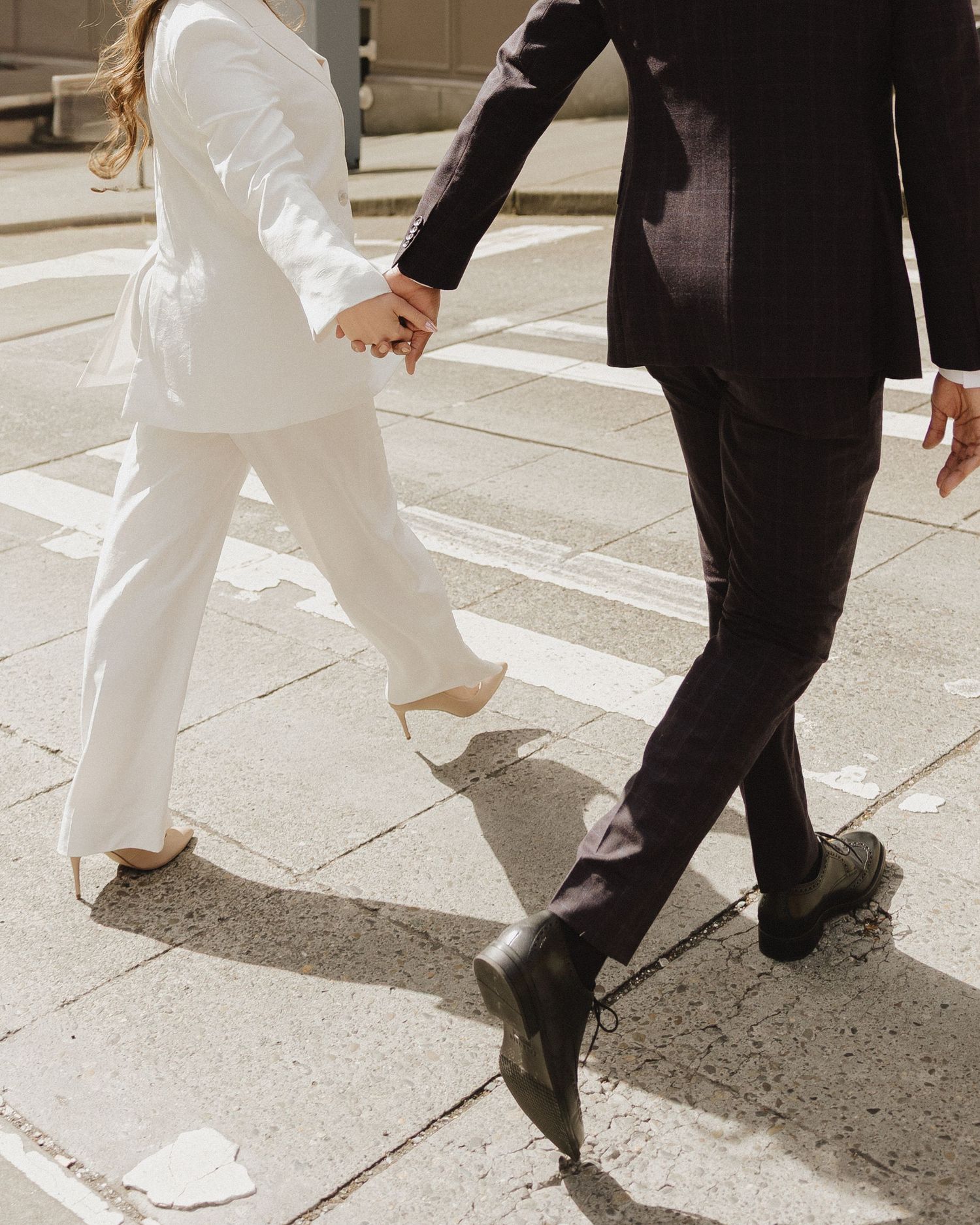 Couple in wedding attire holding hands crossing a street, bride in white suit and groom in dark suit.