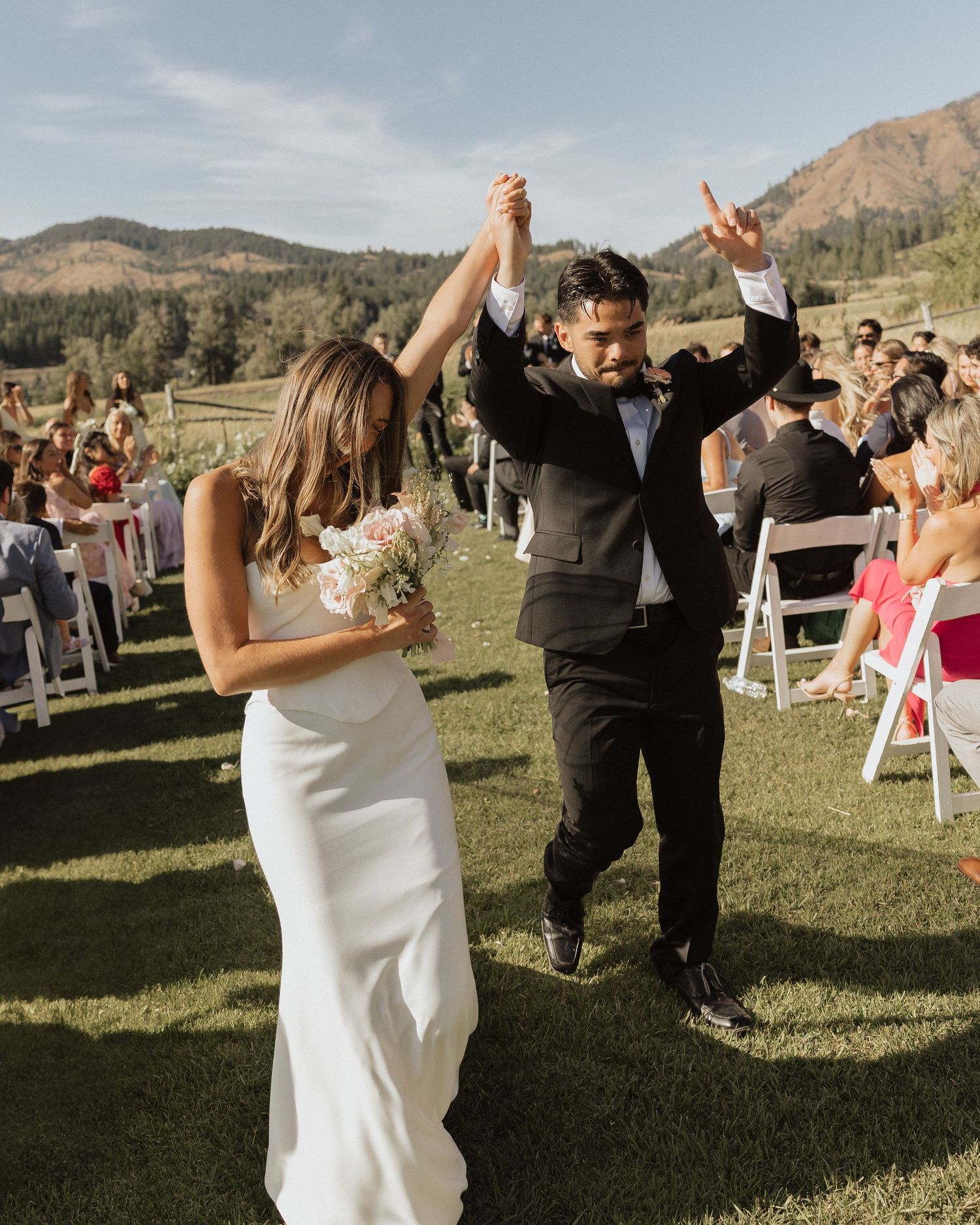 Newlyweds celebrate walking back down aisle at outdoor mountain wedding ceremony on sunny day.