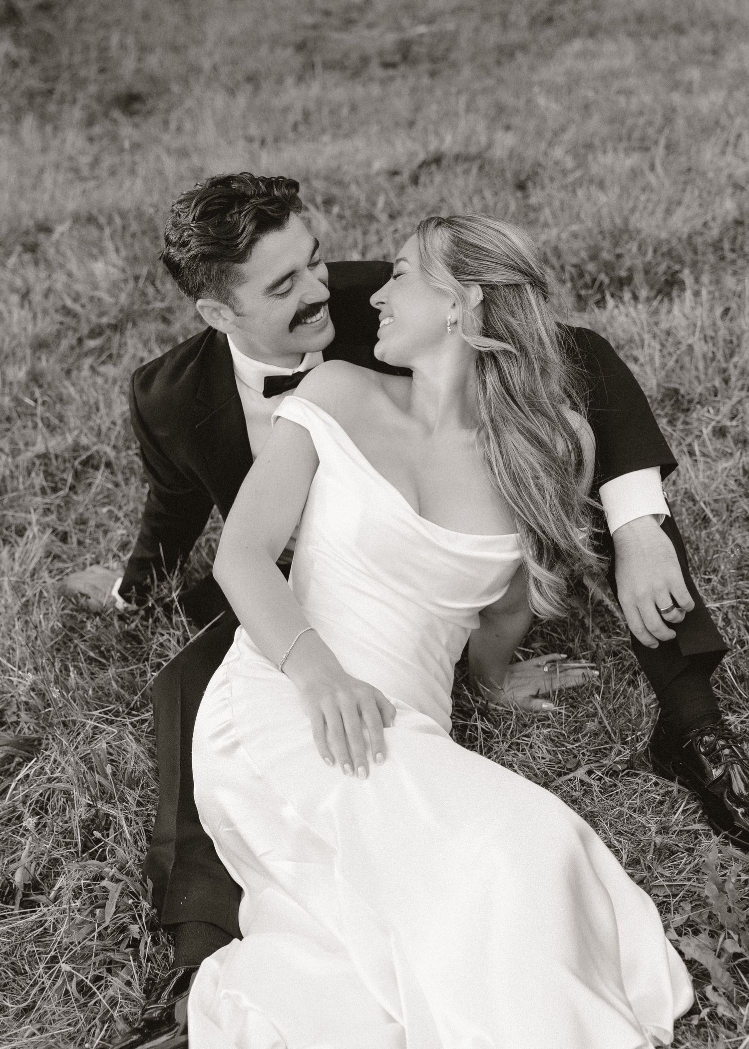 Black and white photo of a bride and groom sitting on grass, gazing at each other lovingly in wedding attire.