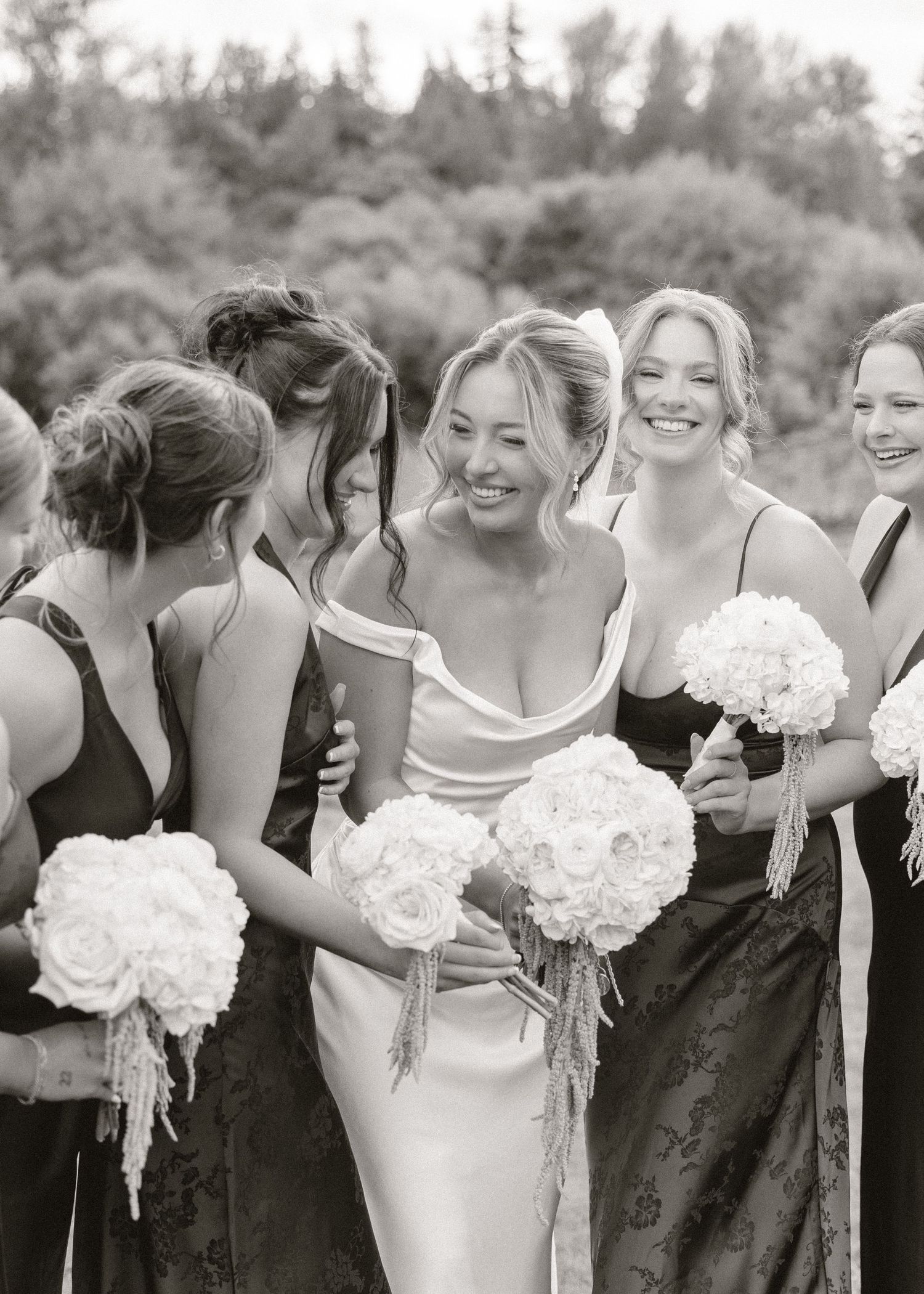 Black and white photo of a smiling bride in a wedding gown surrounded by bridesmaids holding white rose bouquets.