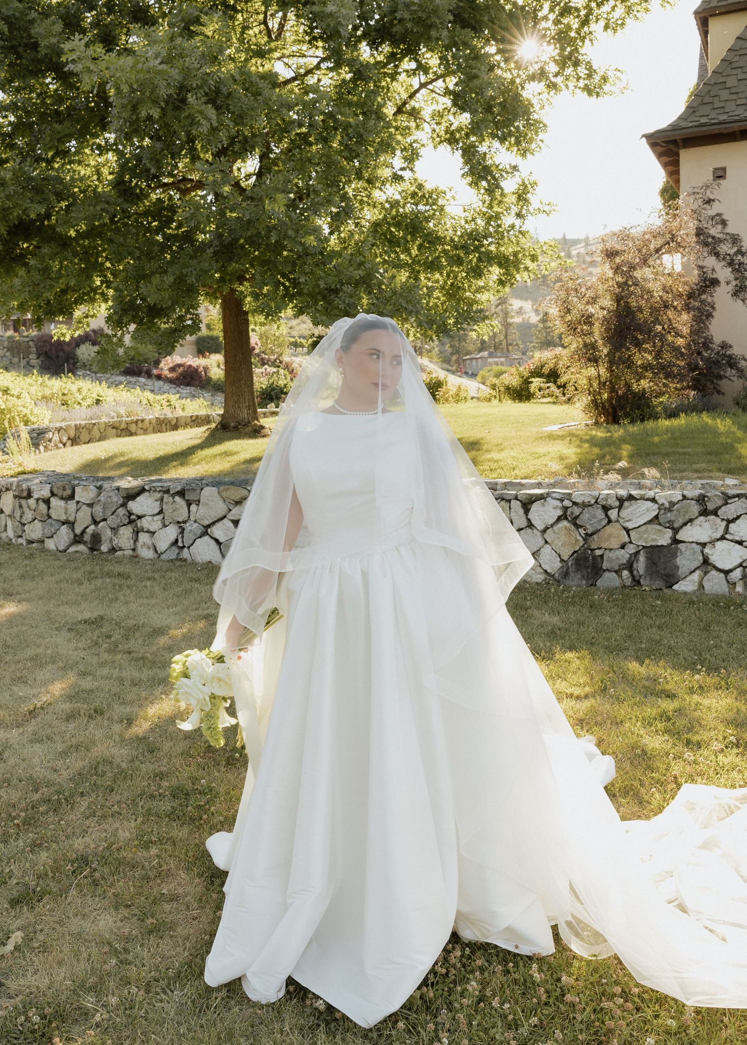 Bride in elegant white wedding gown with veil and bouquet standing on green lawn near stone wall.