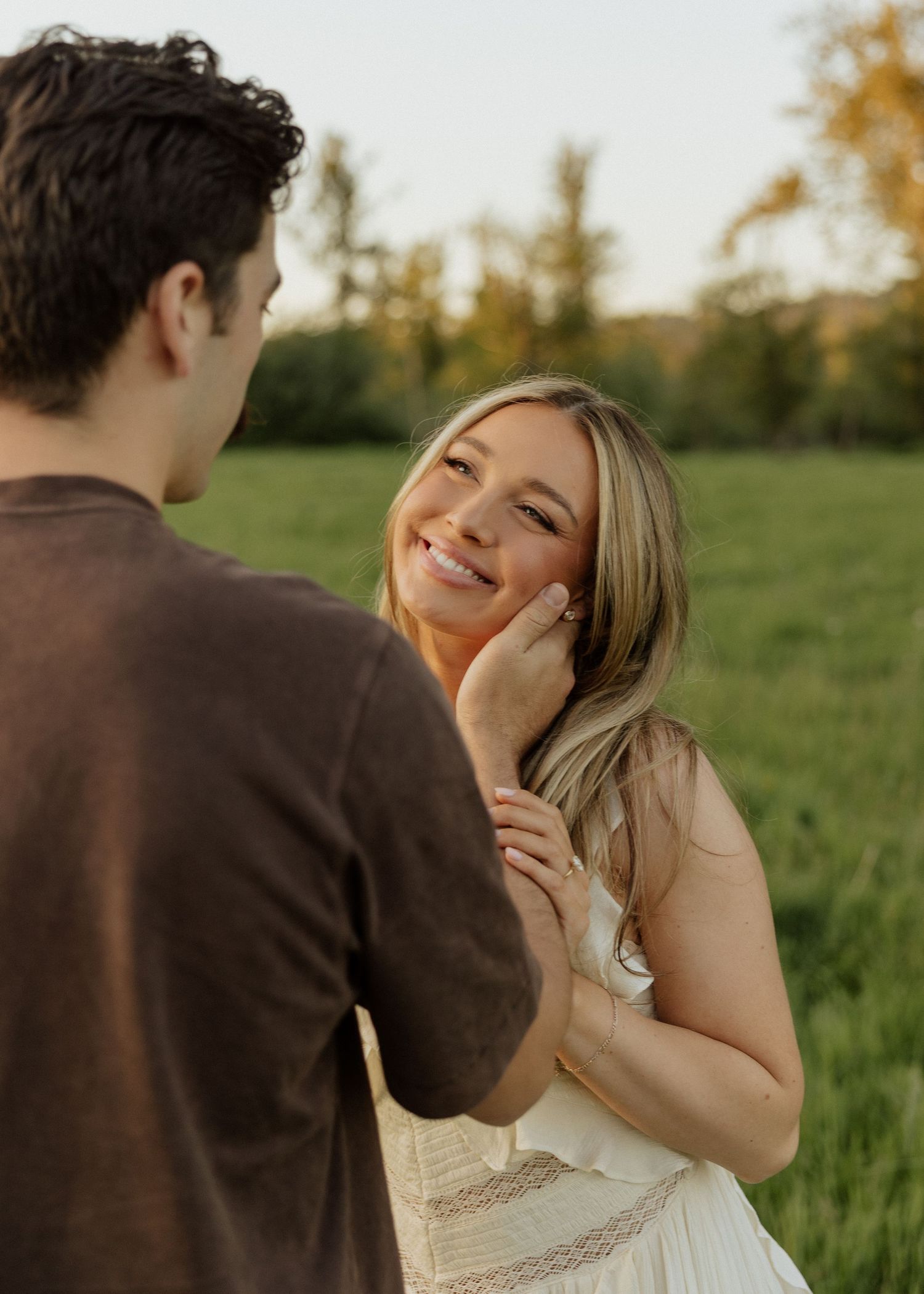 A smiling blonde woman in a white dress gazes at a man in a green field during golden hour.