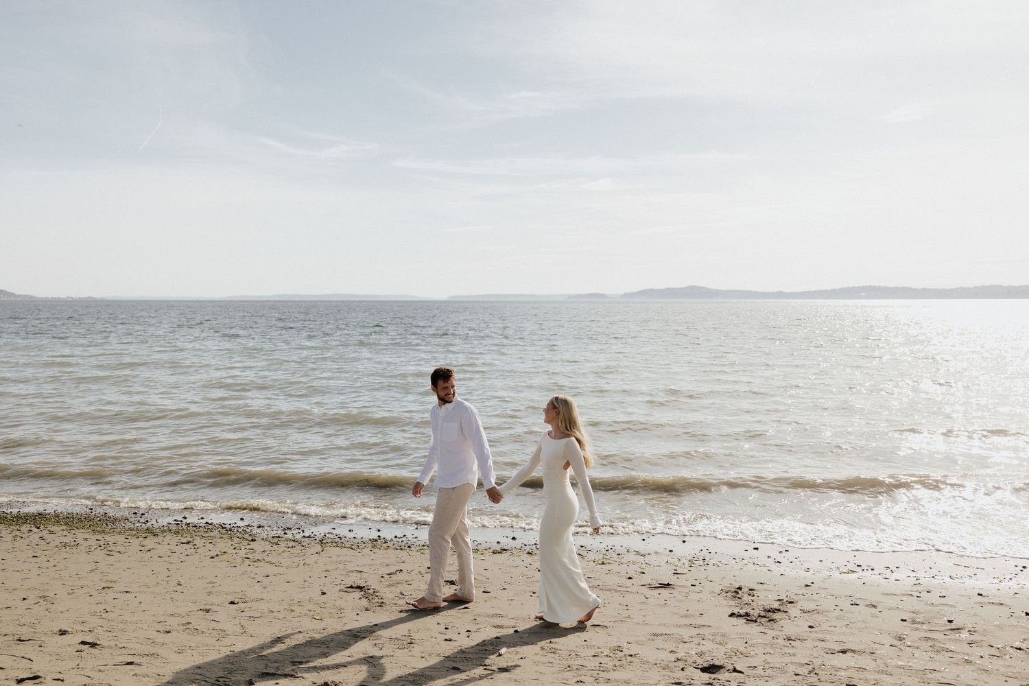 Couple in white outfits holding hands while walking along a sandy beach shore with calm water in the background.